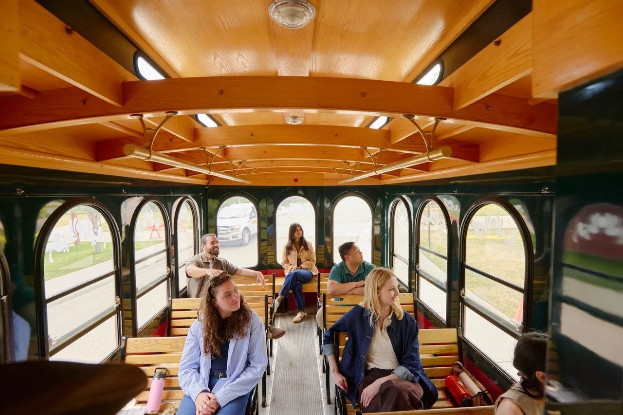 Passengers sit on wooden benches inside a trolley, looking out arched side windows.