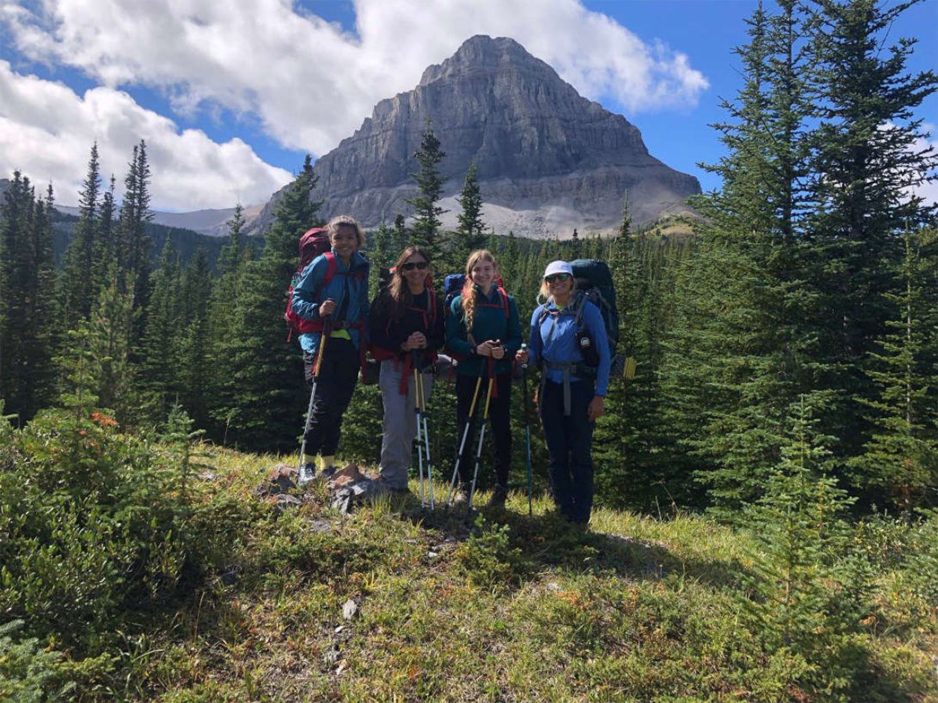 Hikers with backpacks stand among evergreen trees below a rocky mountain peak.