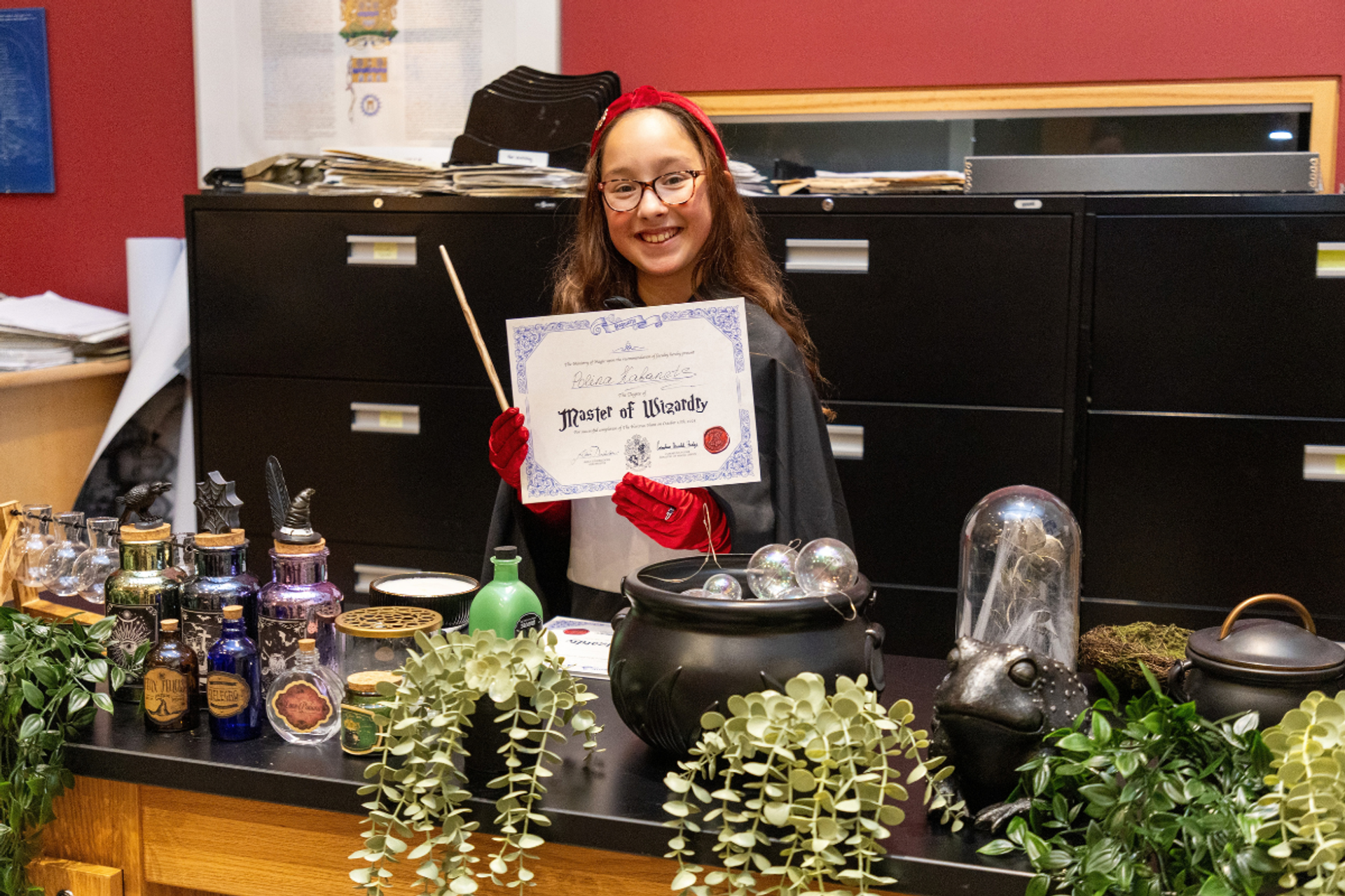 Costumed person holds a Master of Wizardry certificate and wand behind a potion display.