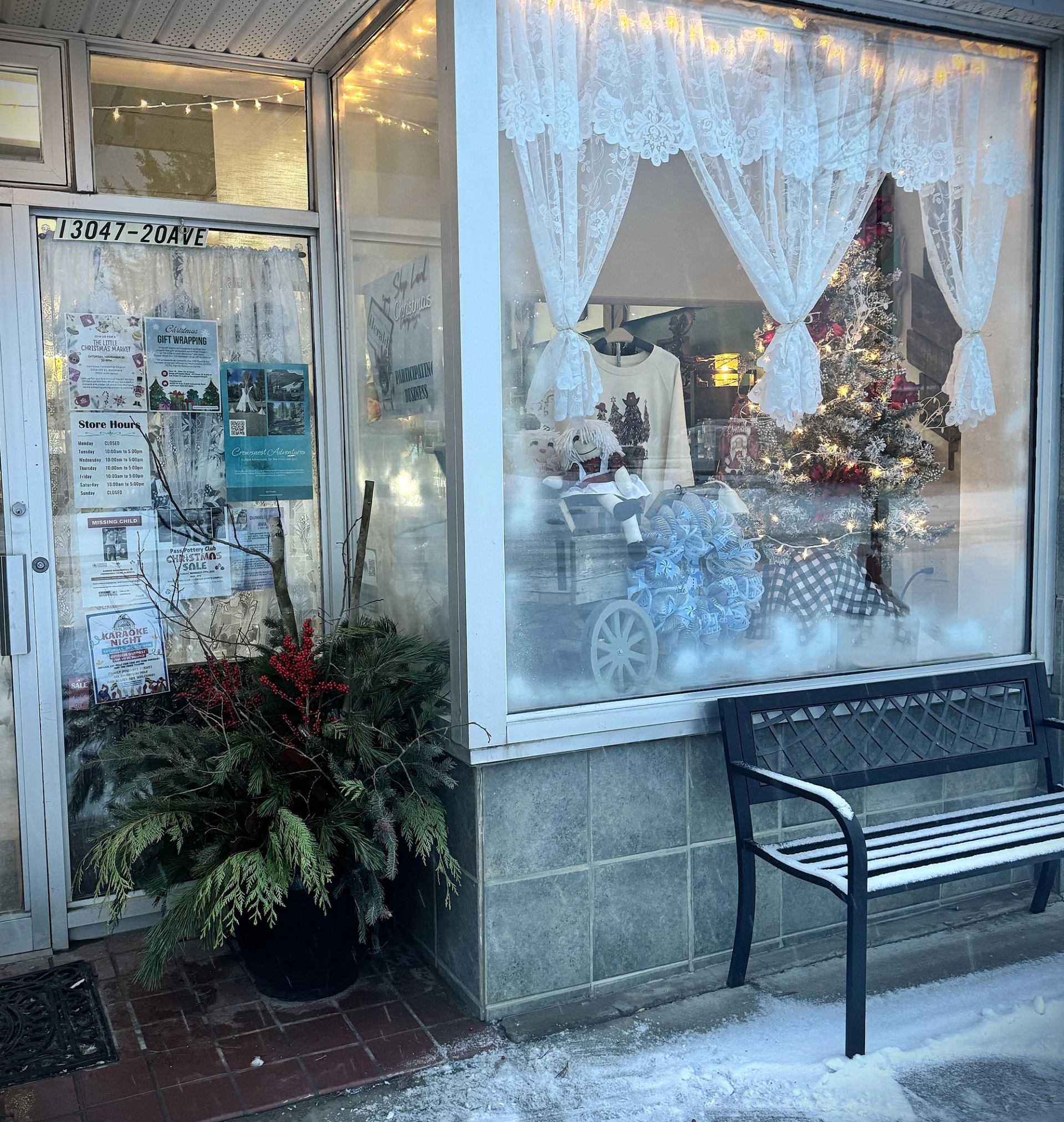 Storefront with lace curtains, holiday décor, greenery arrangement, and a bench beside the entrance.