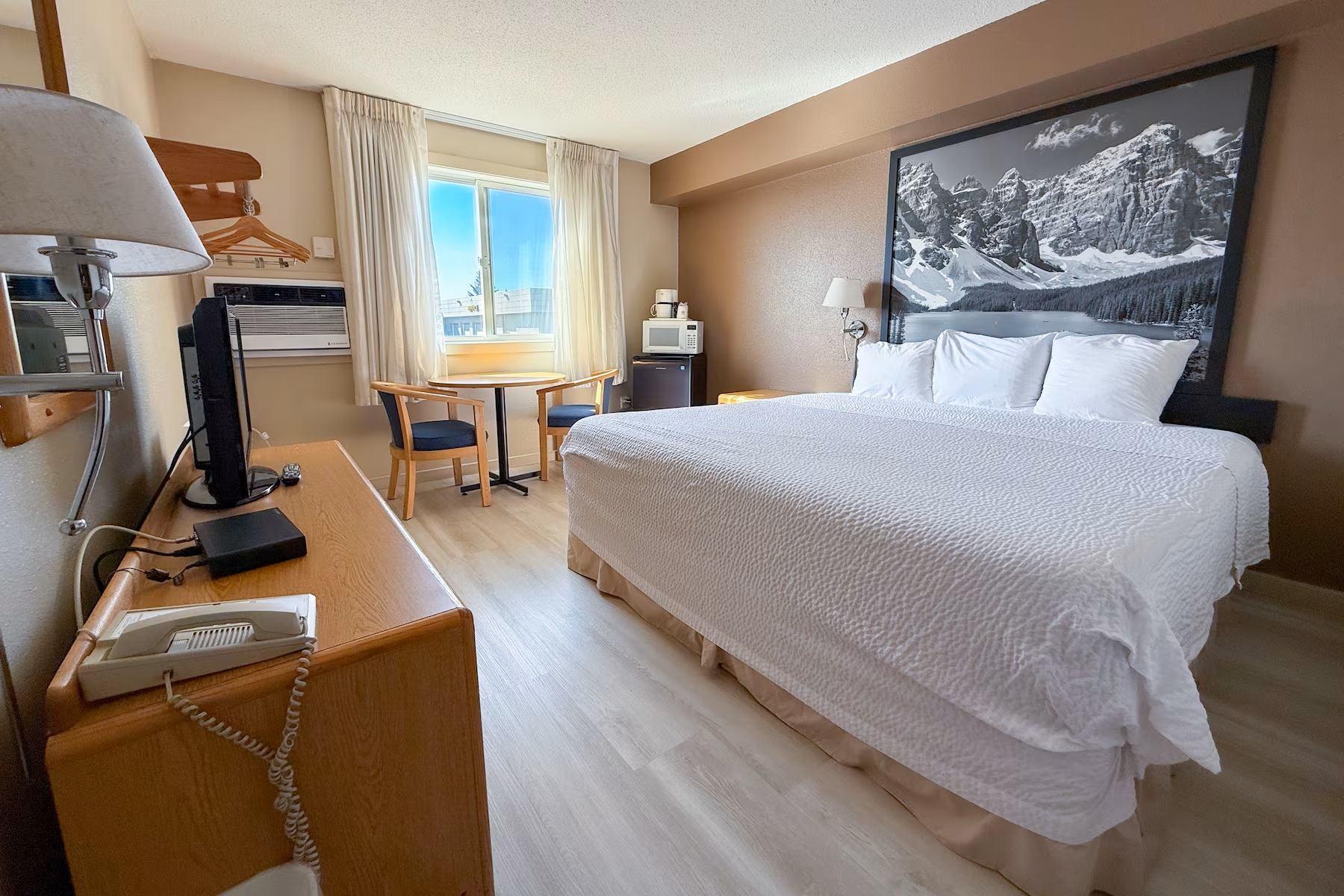Hotel room with king bed, desk, small dining table, and mountain artwork.