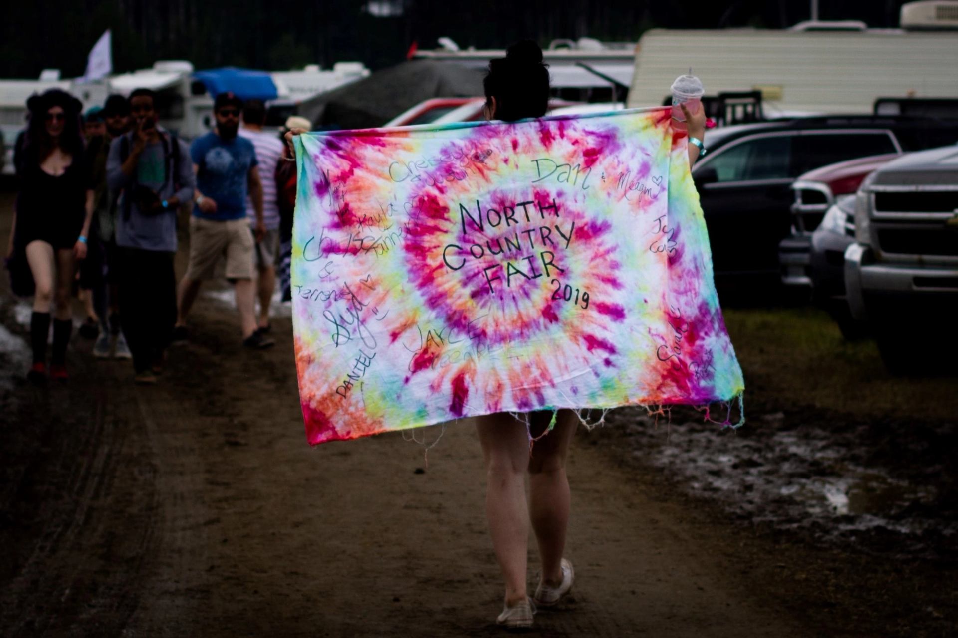 Person walking along a muddy path holding a tie-dye North Country Fair banner with others nearby.