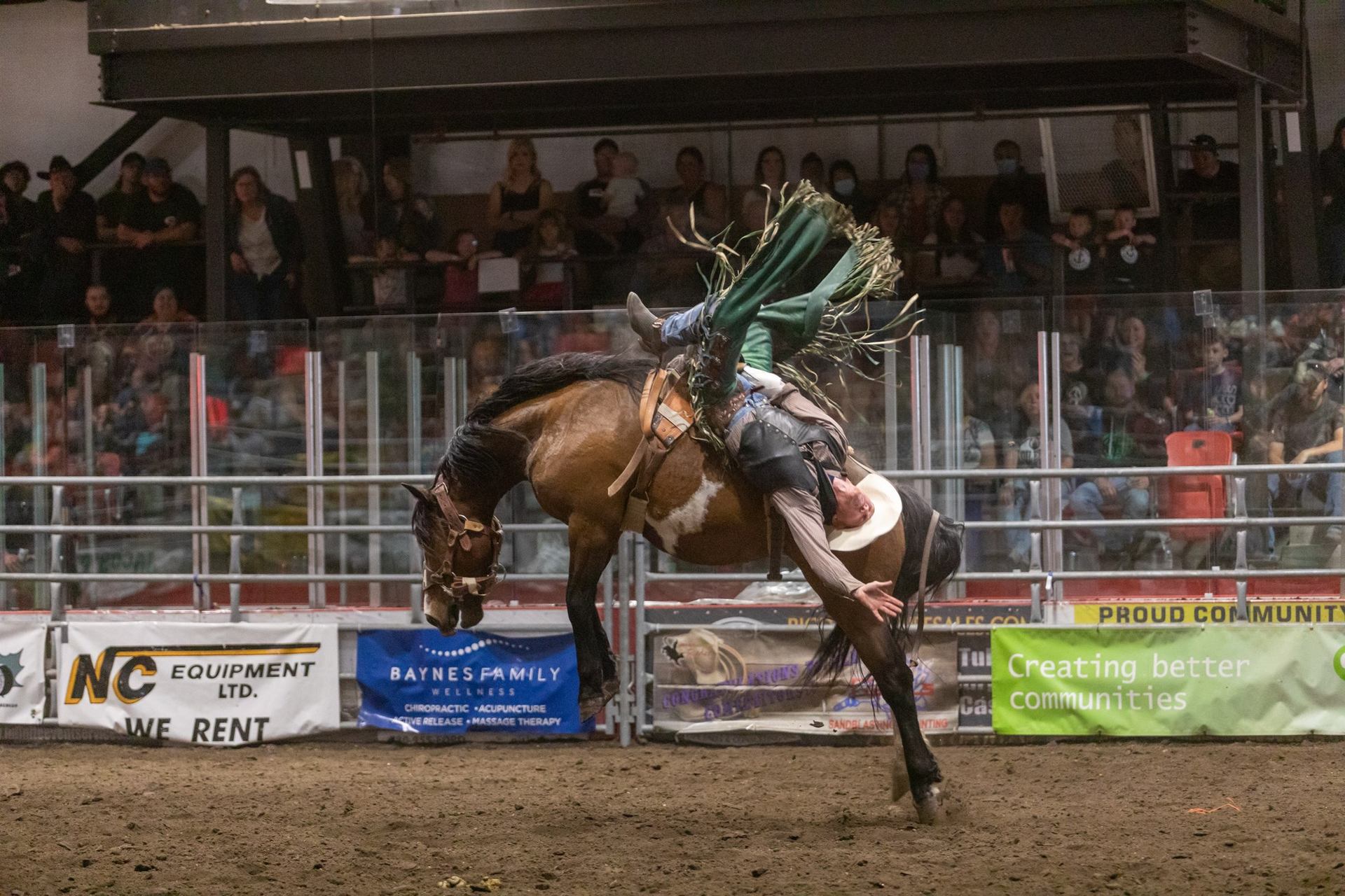 A rider leans back on a bucking horse in an indoor rodeo arena.