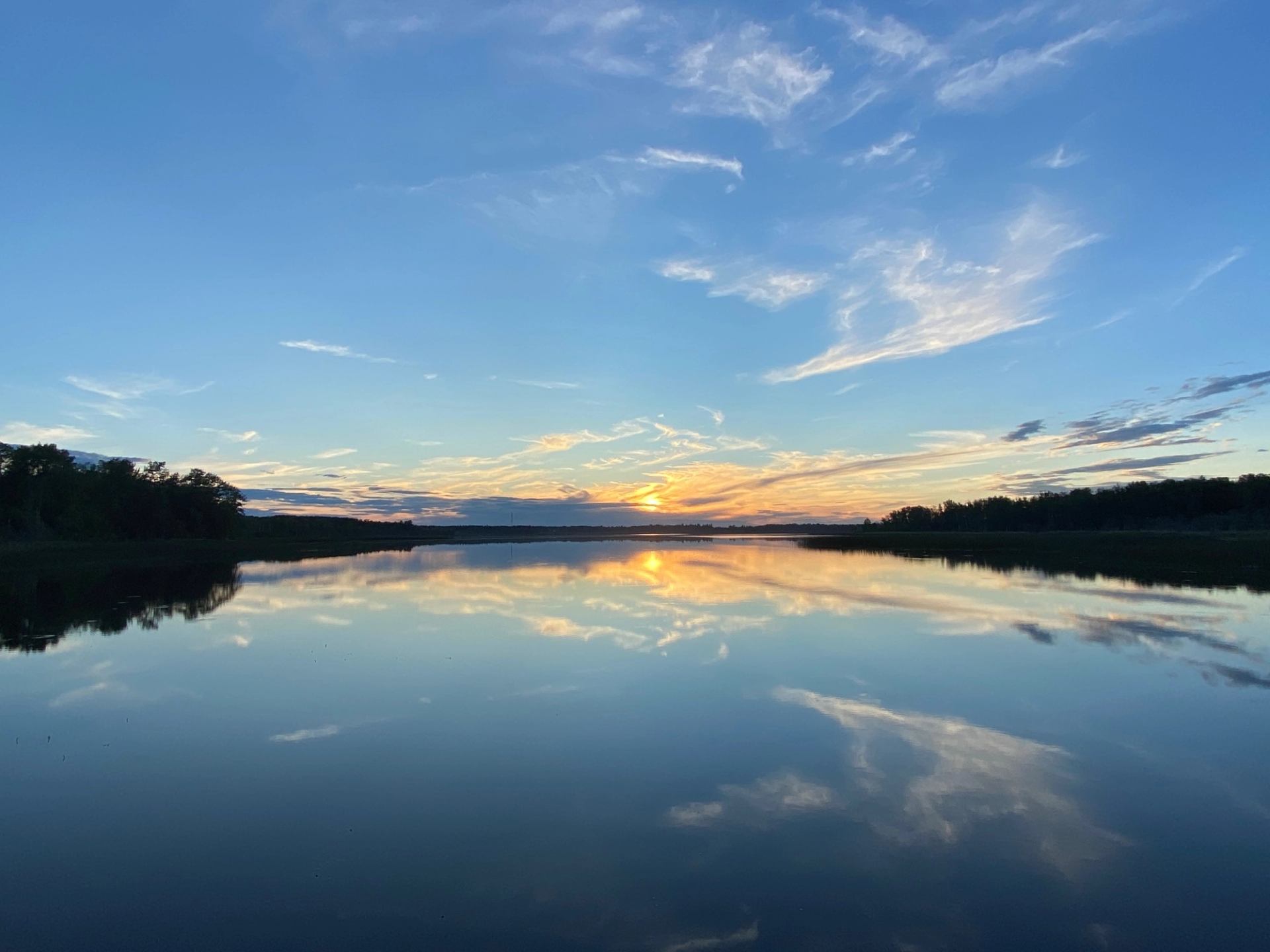 Calm lake reflecting blue sky, scattered clouds and low sun near distant treeline.