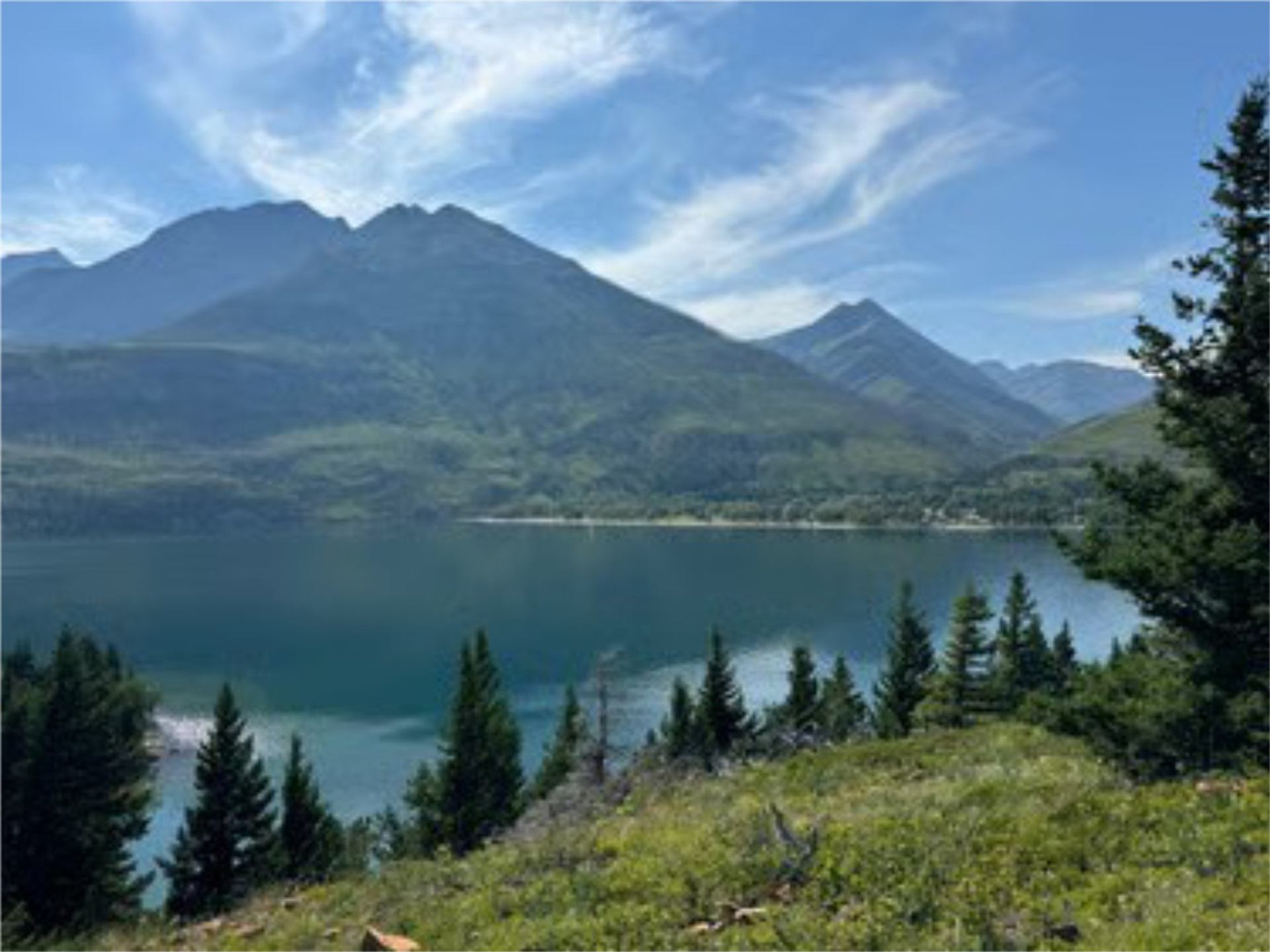 Mountain range and calm lake surrounded by trees under a bright morning sky.