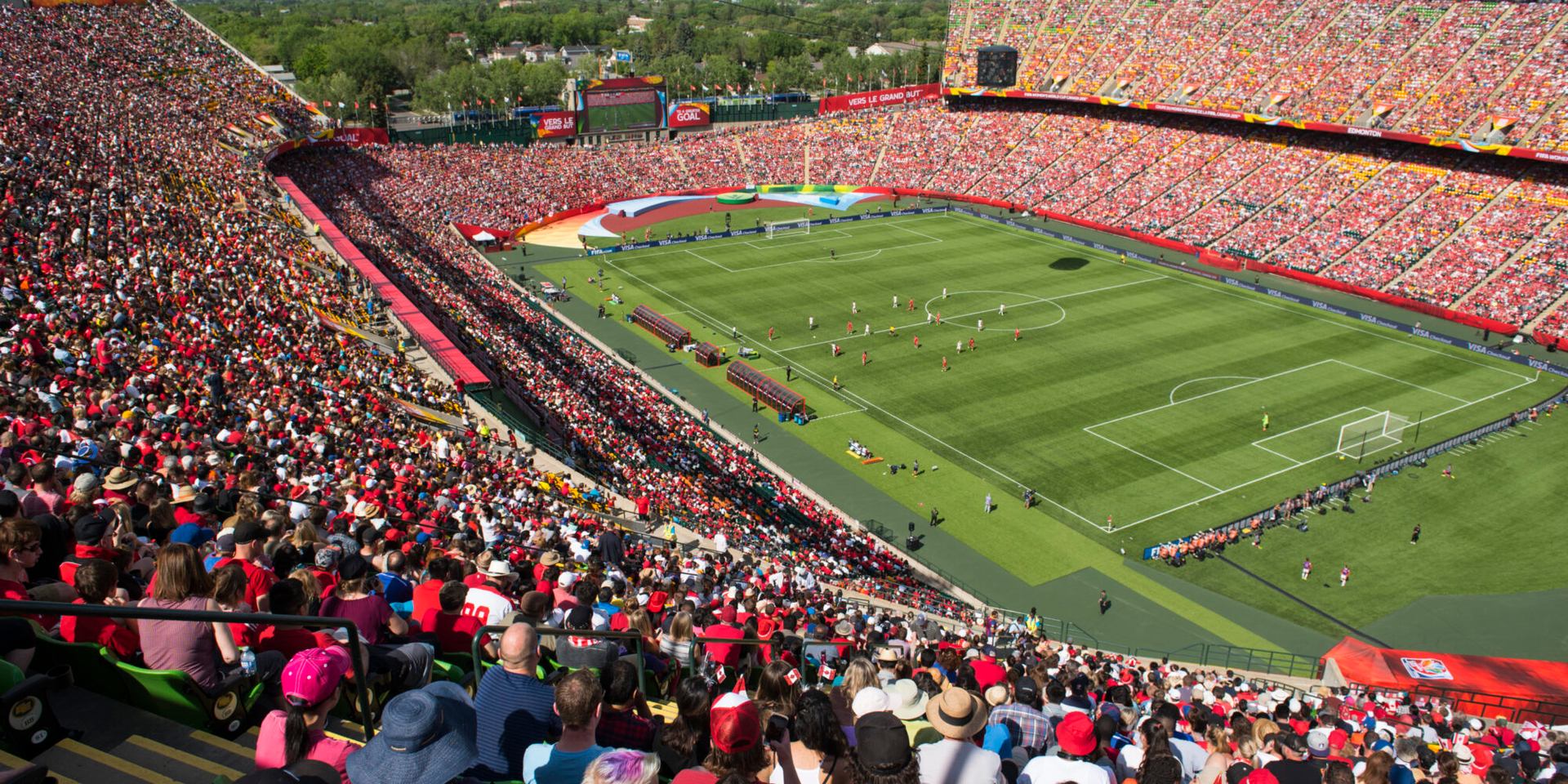 Crowded stadium with fans in red watching a soccer match on green field.