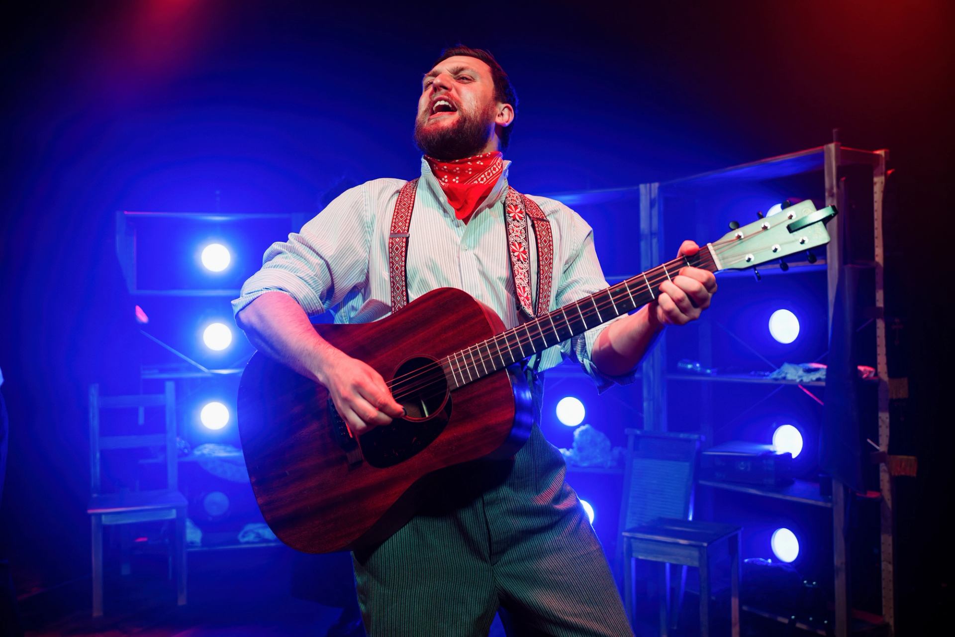 Performer in suspenders plays acoustic guitar on stage under blue and red lights.