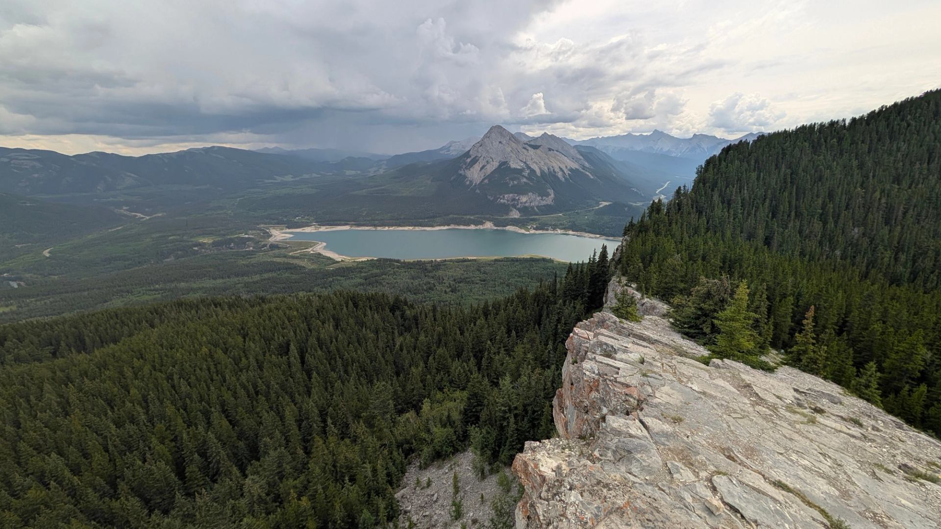 Rocky cliff above dense evergreen forest overlooking lake and distant mountain range.