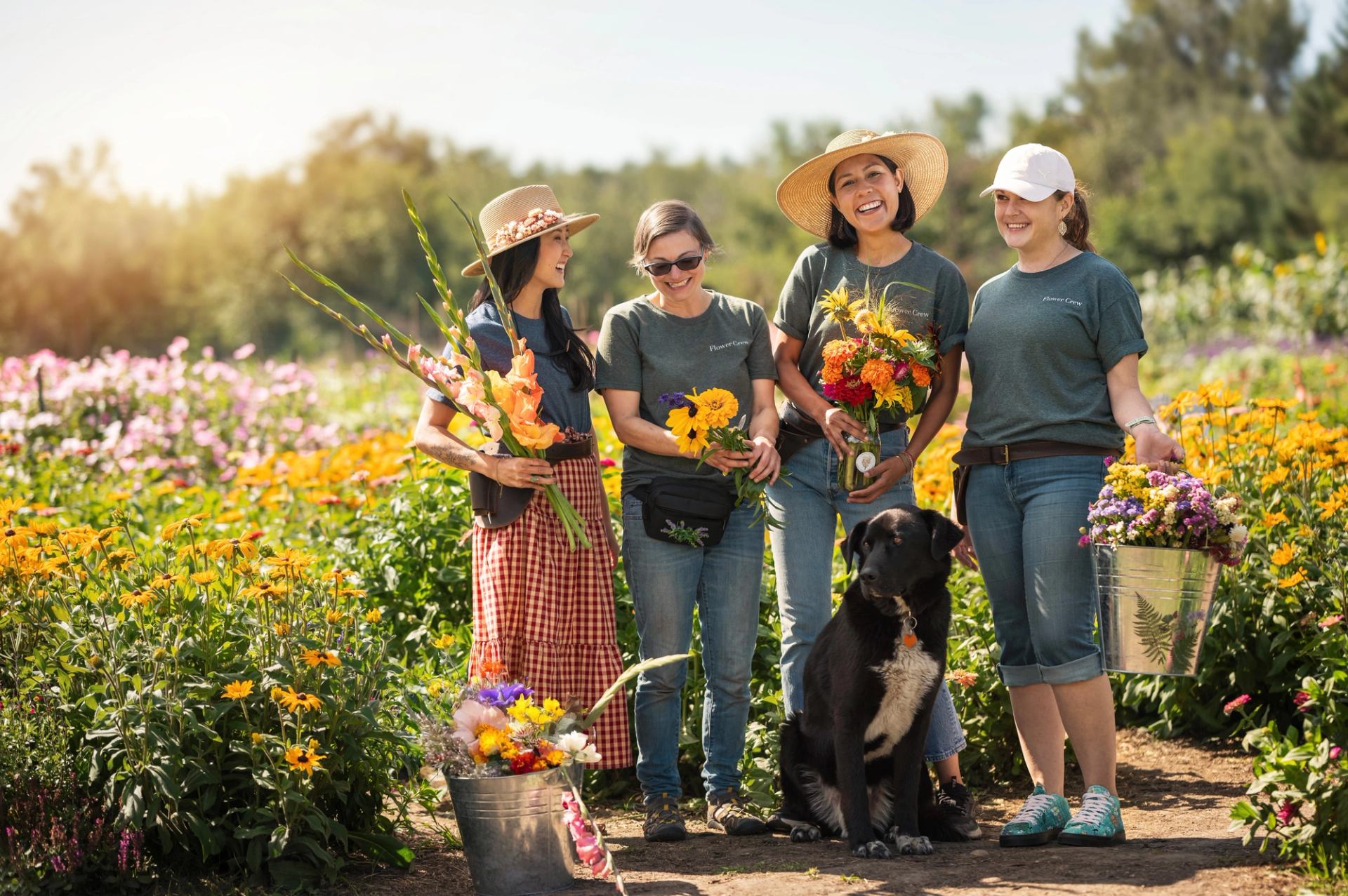 Four people and a dog in a flower field, holding bouquets and baskets of colorful blooms.