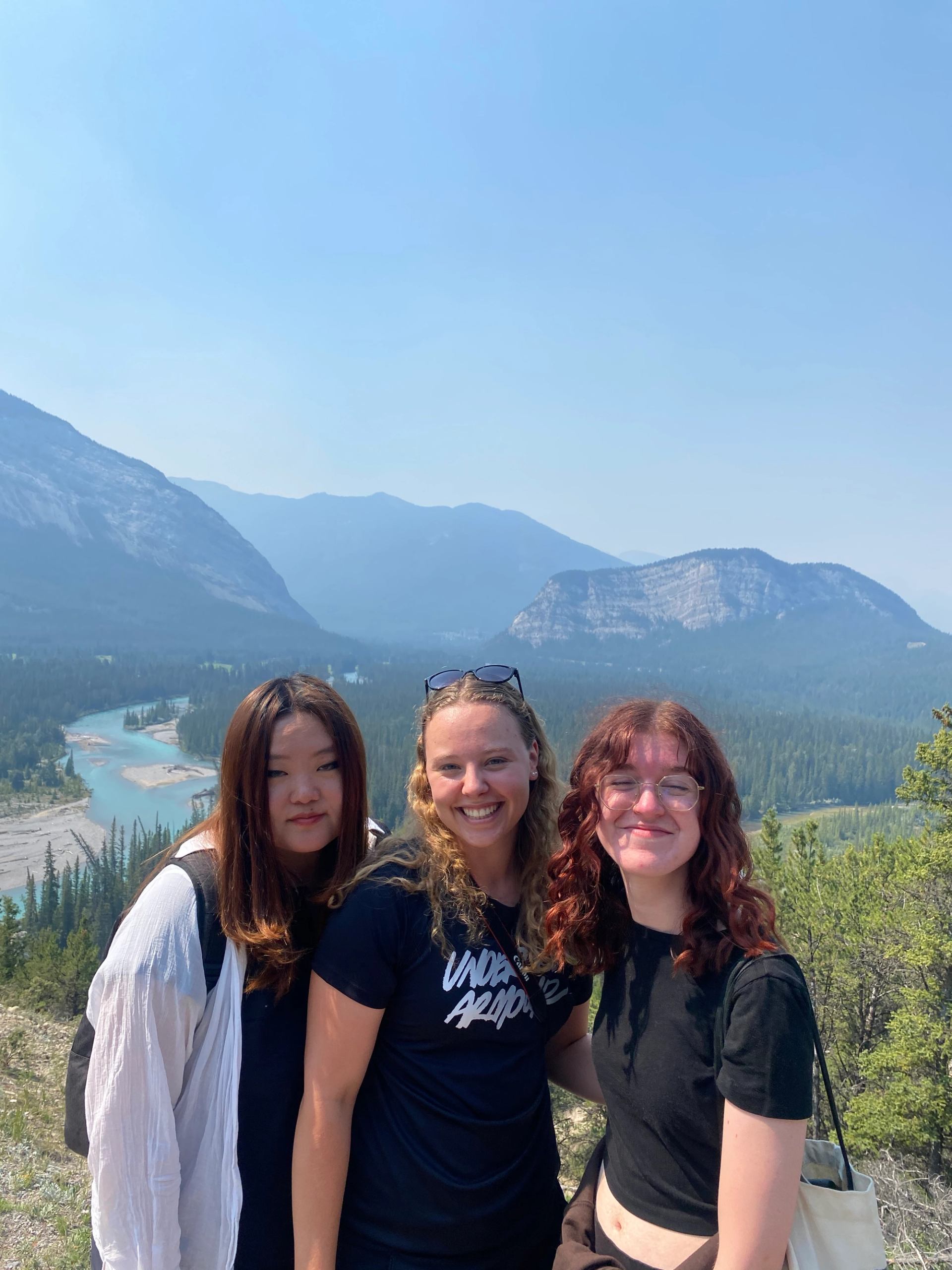 Three friends stand on a viewpoint above forested valley and winding turquoise river.