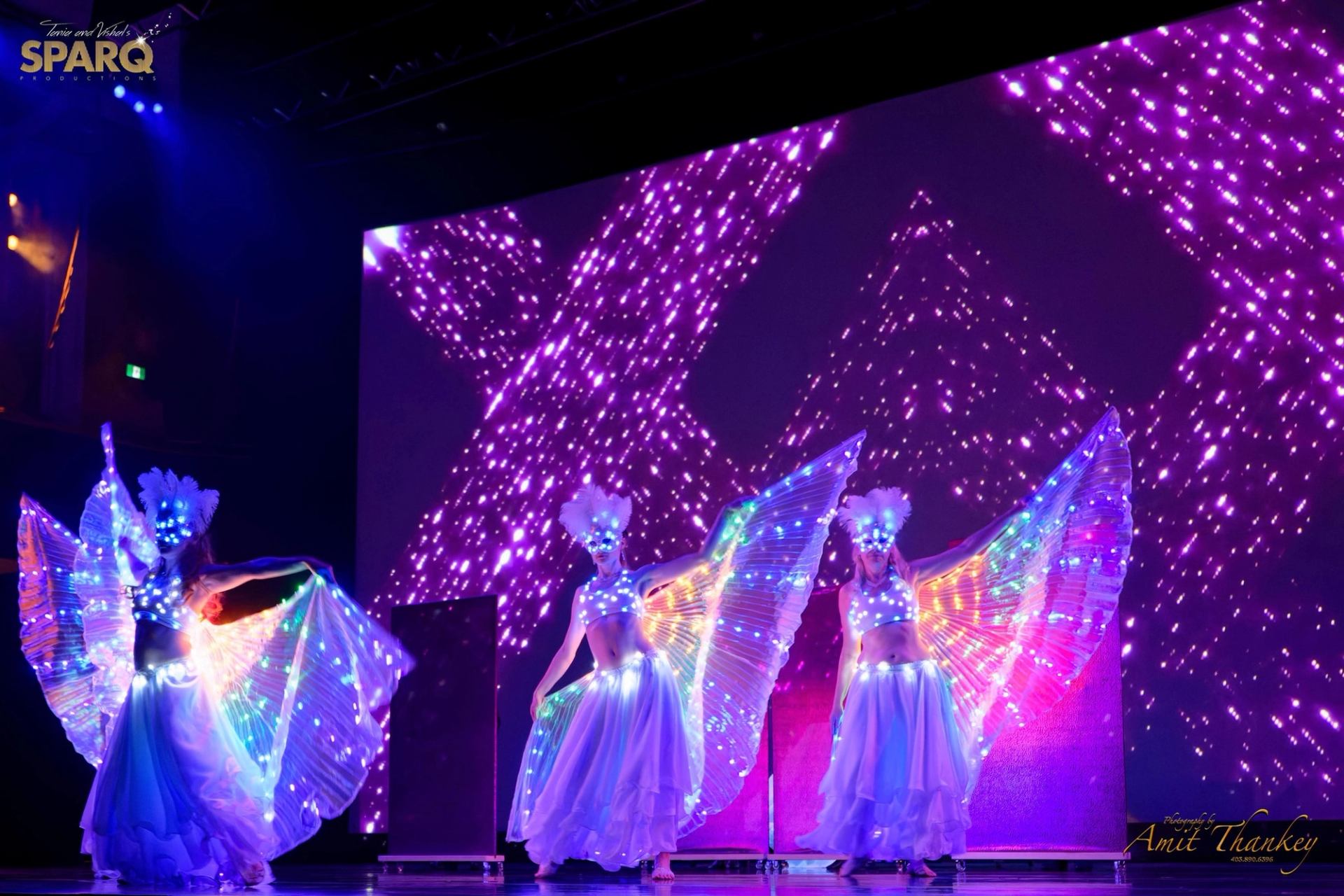 Stage performers in illuminated winged costumes dance before large purple light display.