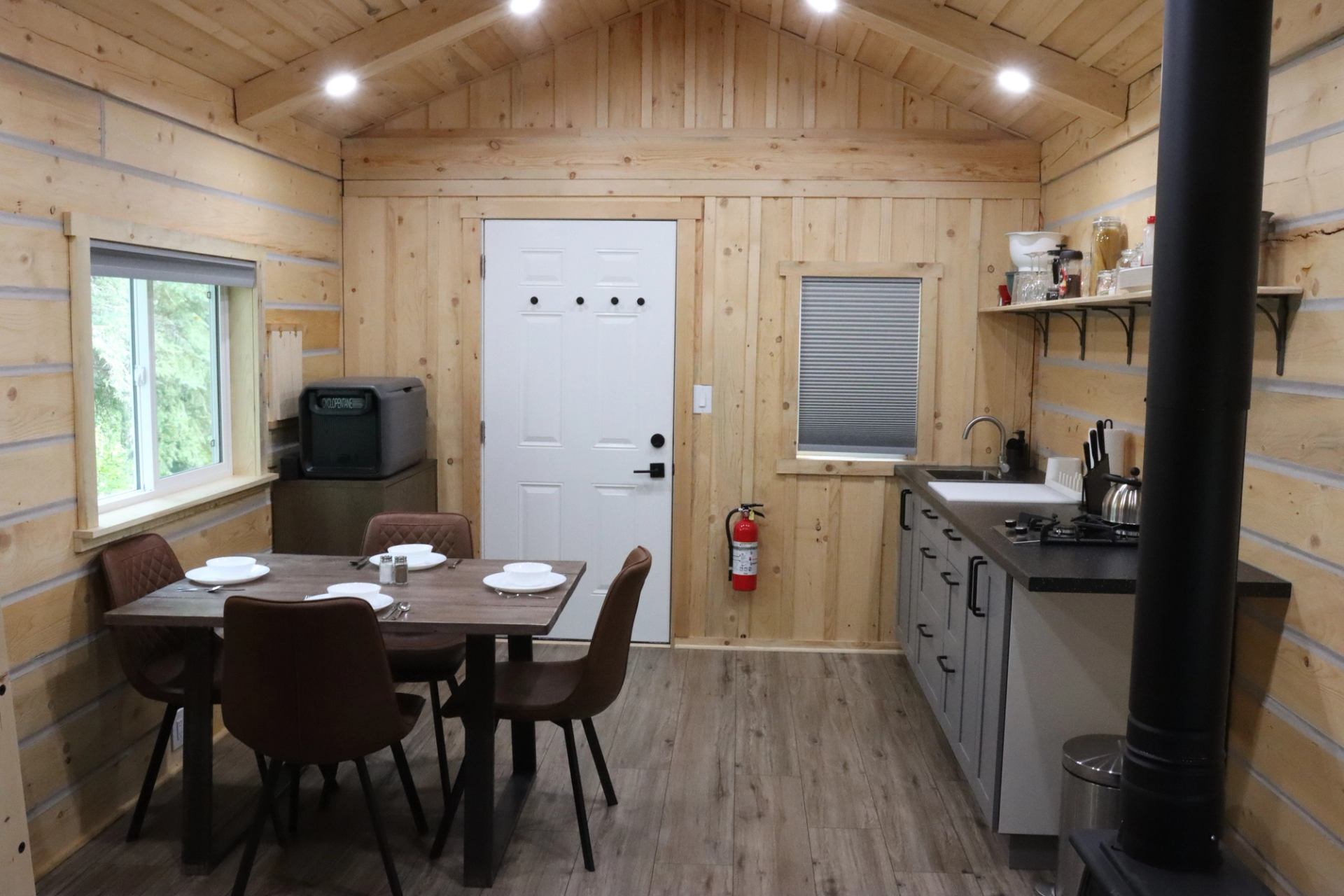 Cabin interior with dining table, kitchen, TV, stove pipe, and door with hooks and extinguisher.