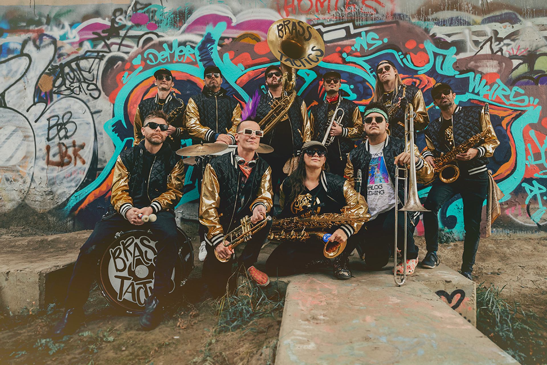 Brass band in matching jackets pose with instruments before colourful graffiti-covered wall.