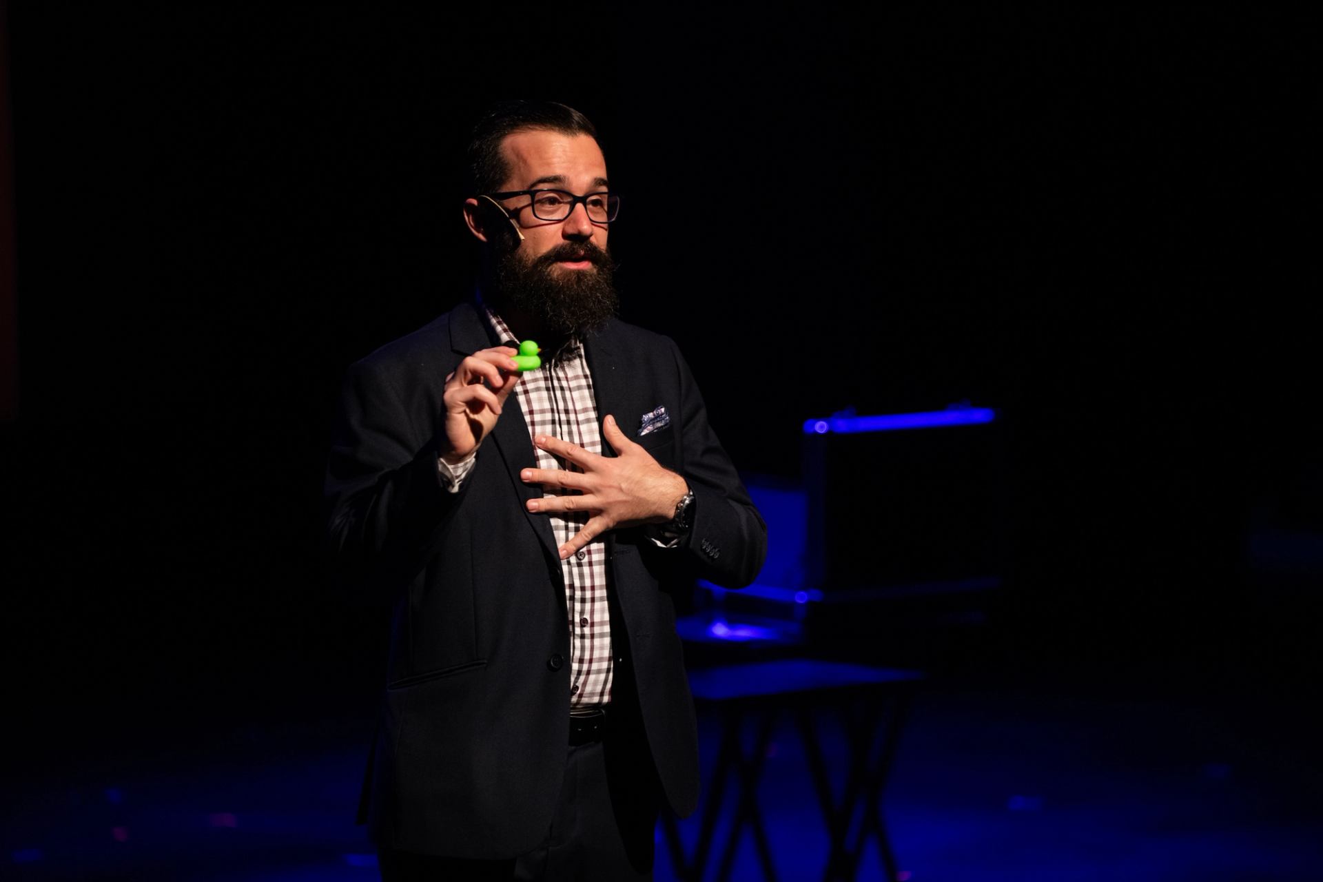 Speaker on dark stage holding small green object and gesturing with other hand.