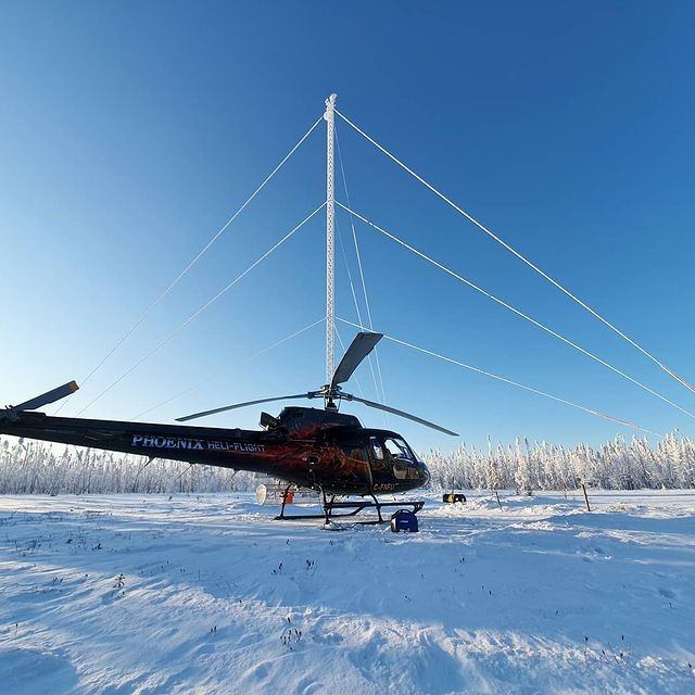 Black and red helicopter on snowy ground near tall power line structure under clear blue sky.