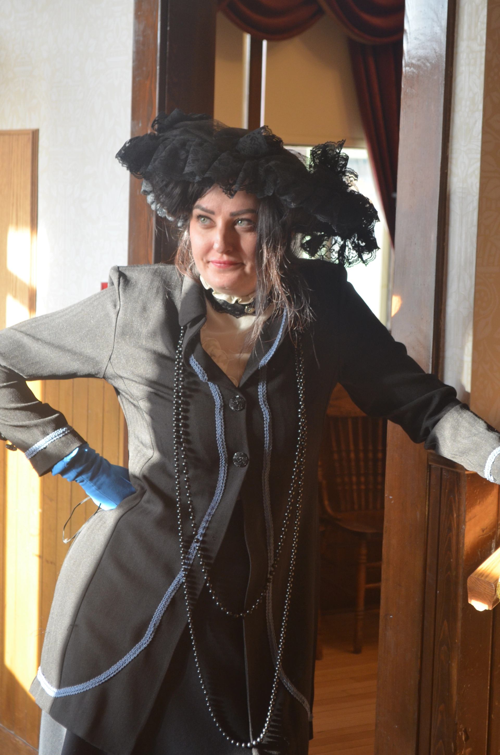 Person in a black period costume and lace hat leans against a wooden doorway.