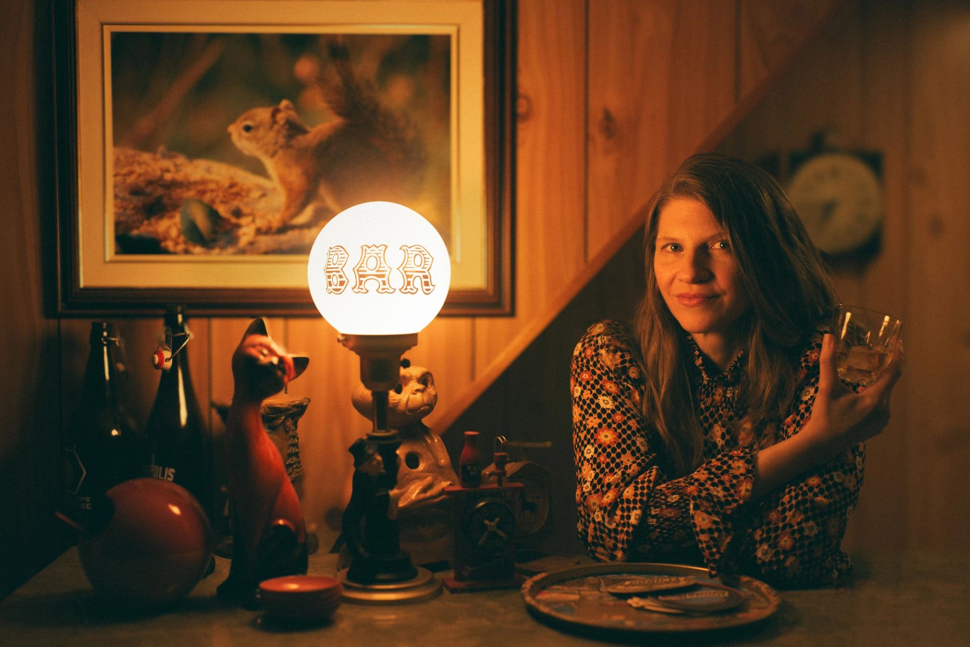 Woman sitting at retro bar, holding drink beside glowing BAR lamp and squirrel painting.