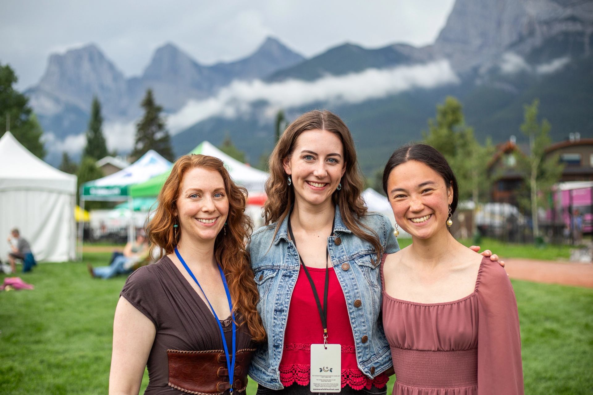 Three people stand together at an outdoor event with tents and mountains behind them.