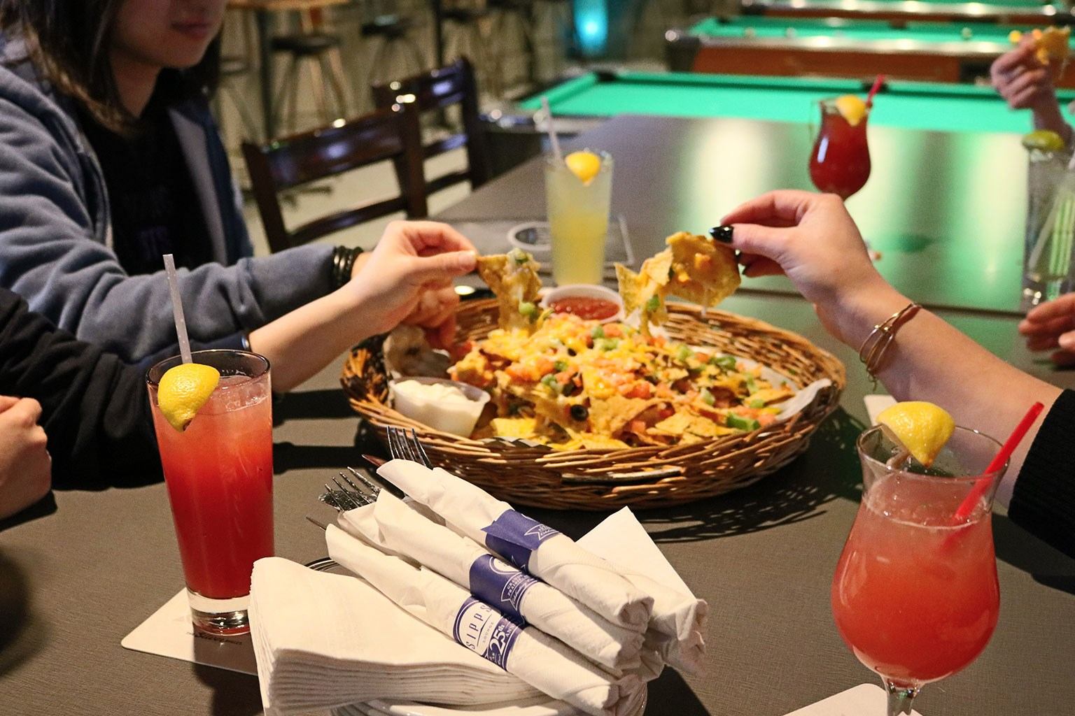 Friends sharing nachos and cocktails at a lively bar with a pool table.