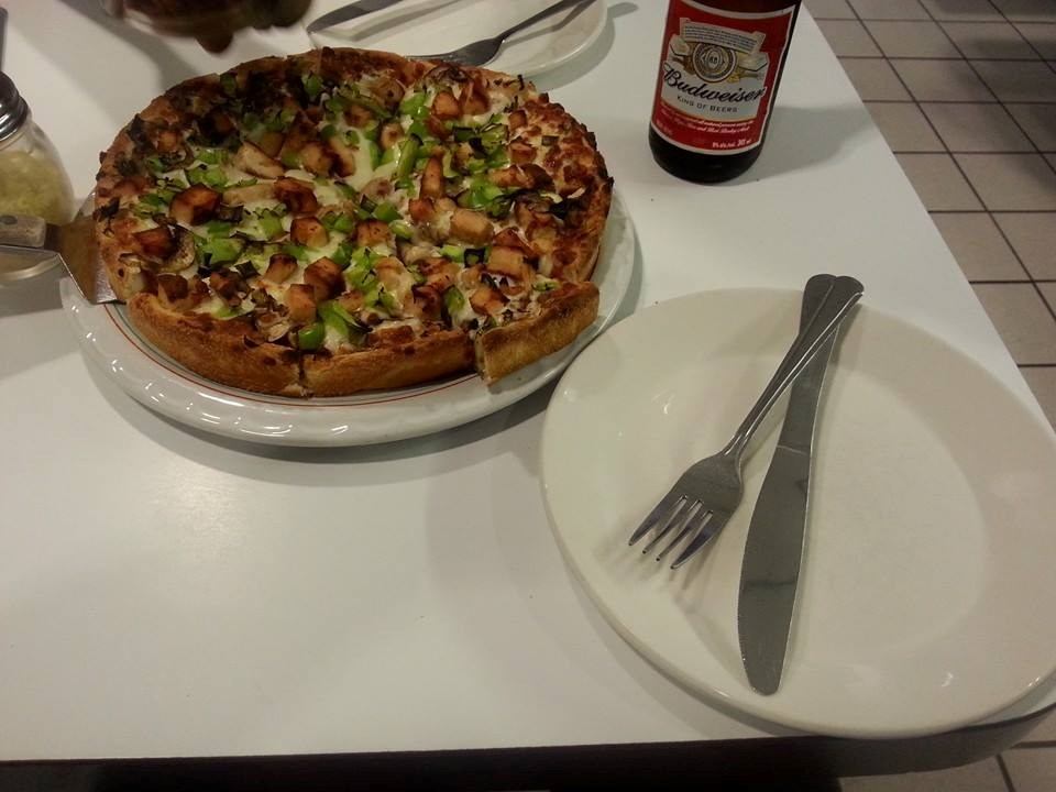 Pizza on plate beside Budweiser bottle and empty plate with knife and fork.
