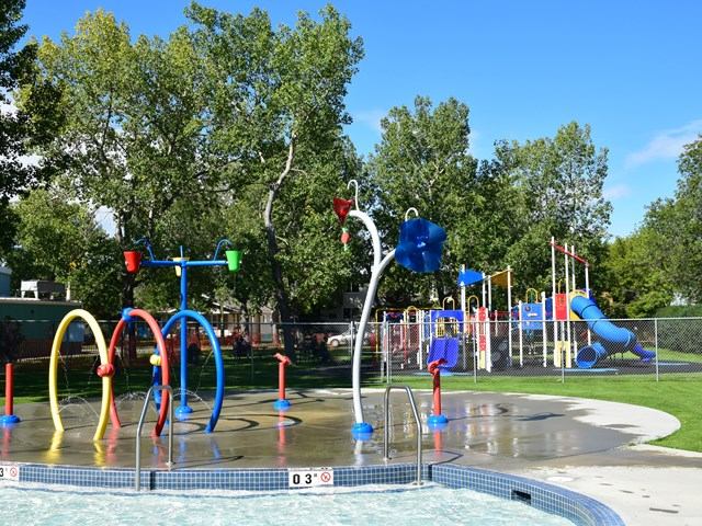 Outdoor splash park with water sprayers, tipping buckets, and playground in the background.
