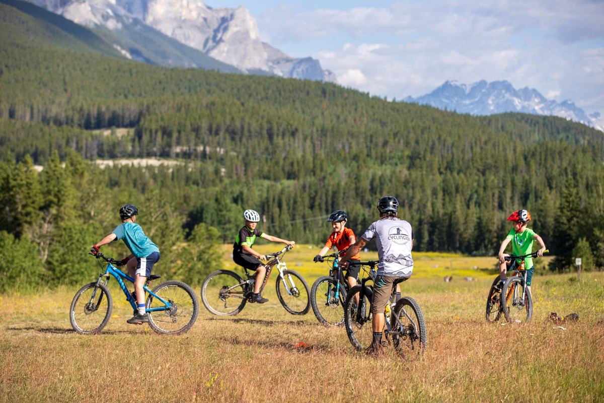 Five cyclists wearing helmets stand on grassy field surrounded by lush green forests and distant mountains under clear blue sky.