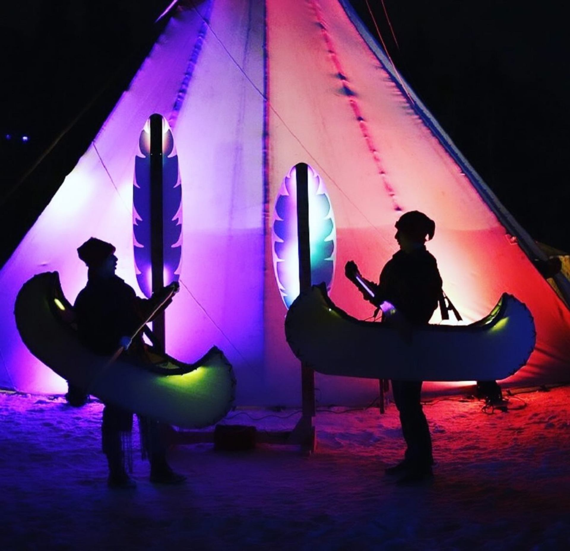 Two people holding glowing canoes in front of a brightly lit tipi at Flying Canoë Volant festival.