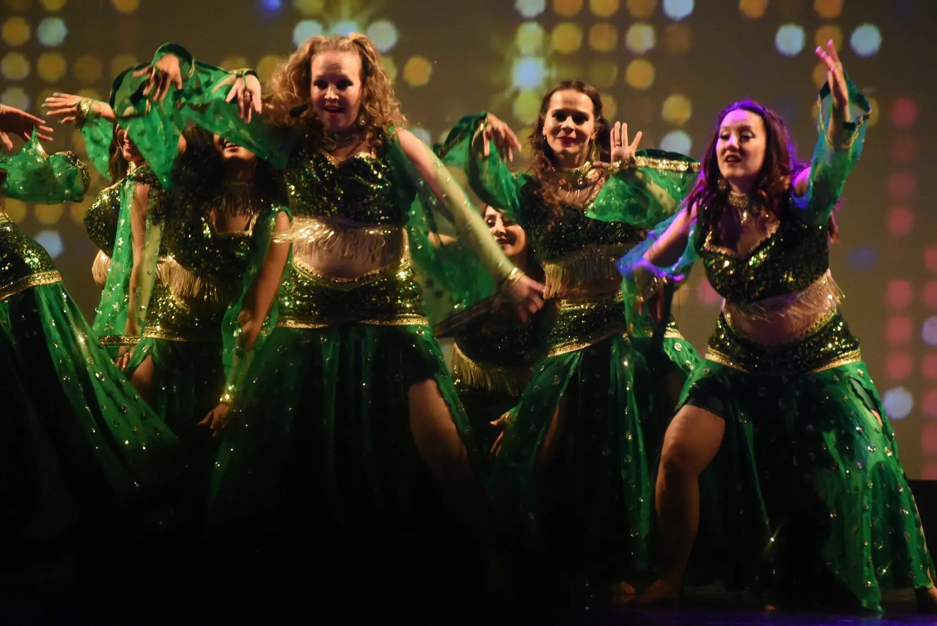 A group of female dancers in sparkling green costumes and gold accents performing a synchronized dance on stage, with dramatic lighting and a colorful bokeh-style background.