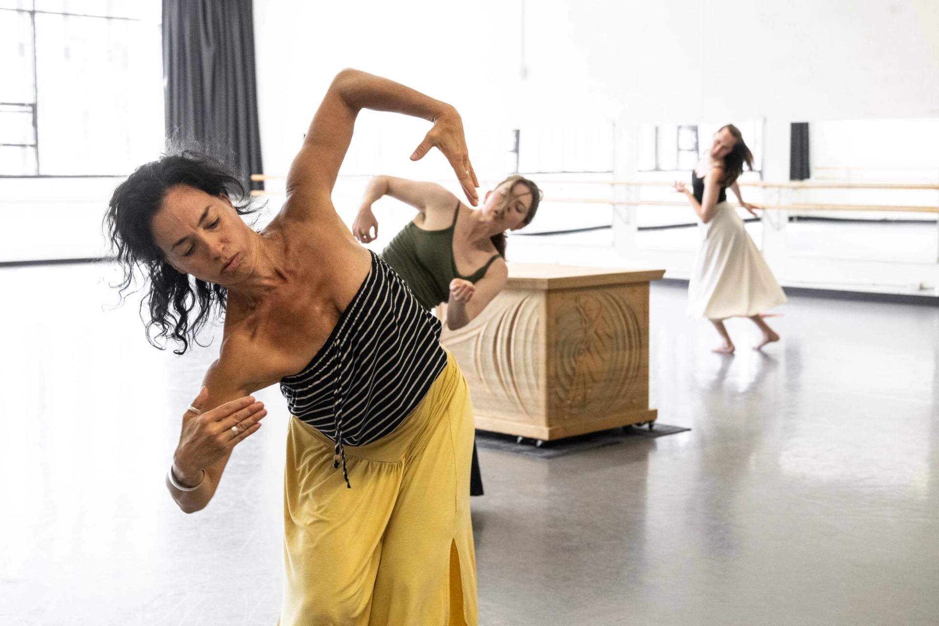Contemporary dancers bend and reach in a bright studio beside a carved wooden box.