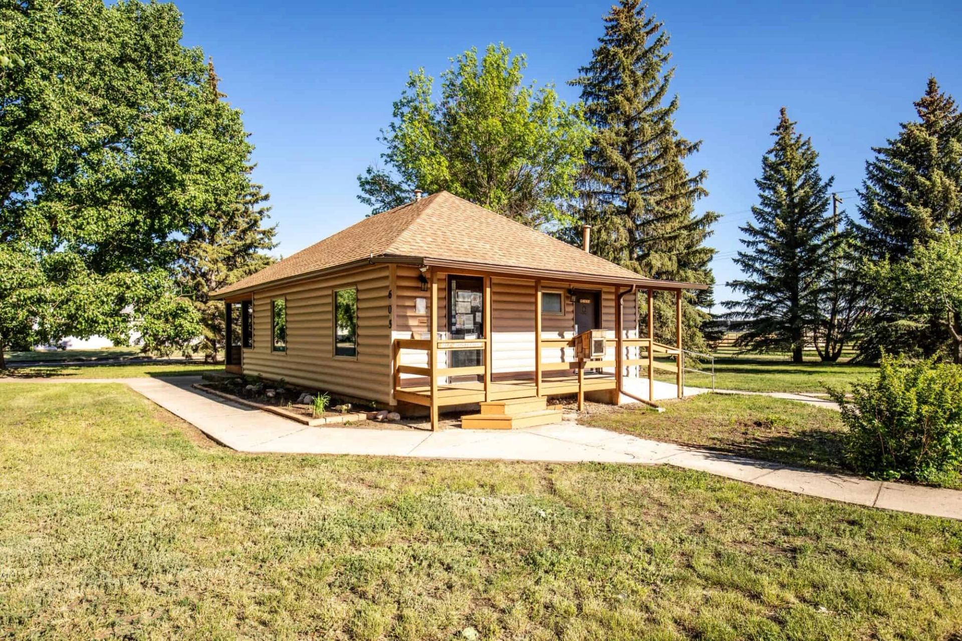 Small tan cabin with covered porch, concrete paths and surrounding tall evergreen trees.