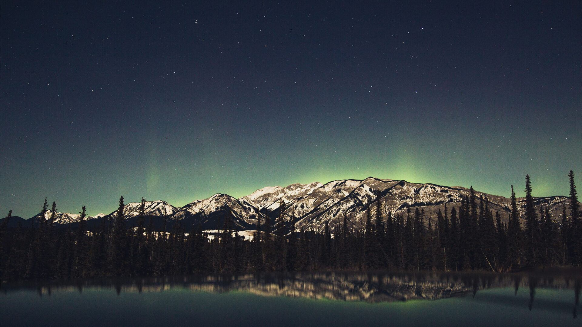 Snowy mountains and trees reflected in calm water under faint green aurora and a starry sky.