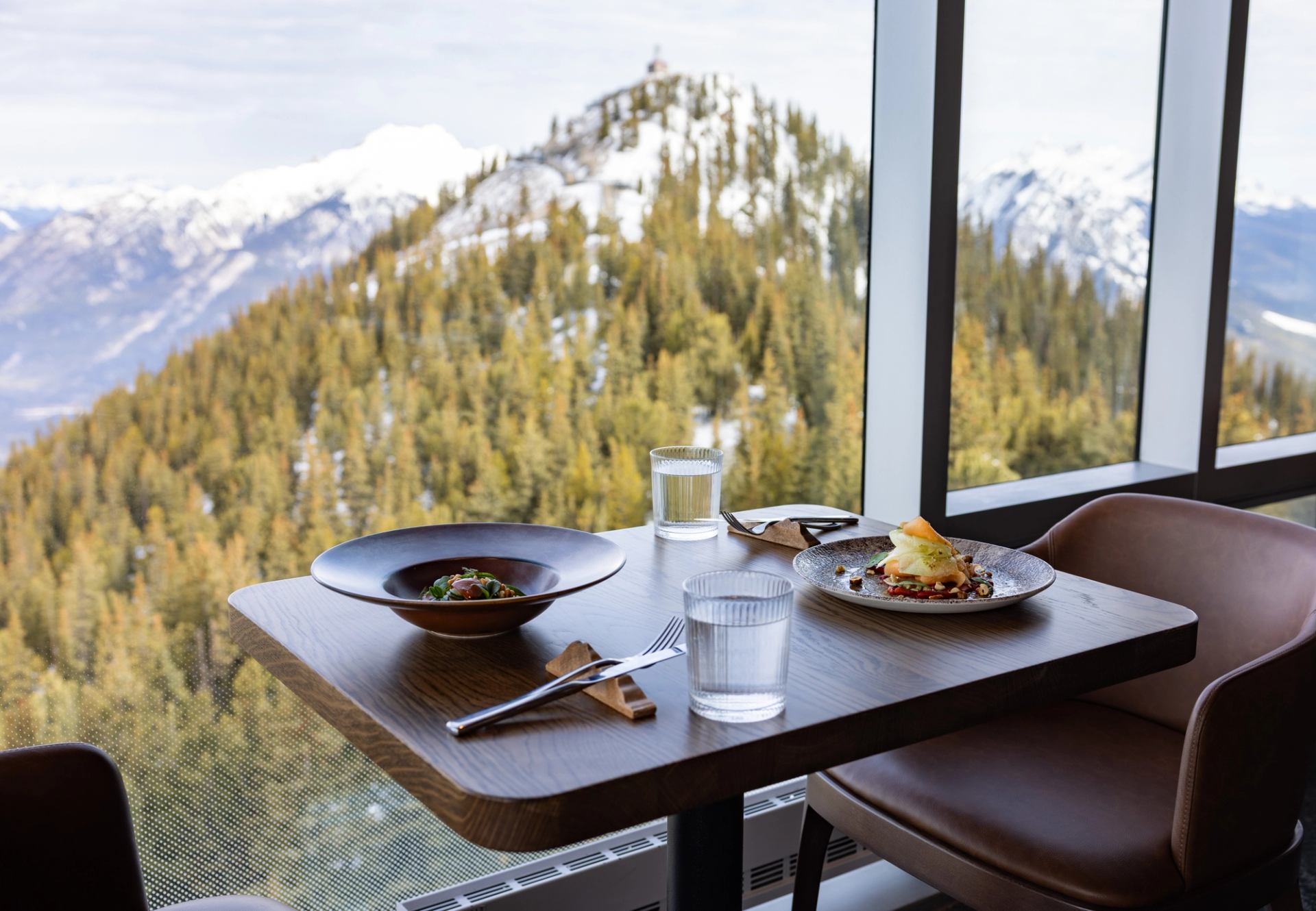 Restaurant table with plated meals and water glasses beside large window overlooking snowy mountains.