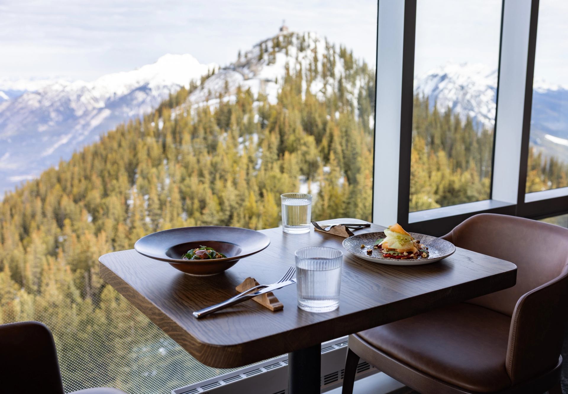 Restaurant table with plated meals and water glasses beside large window overlooking snowy mountains.