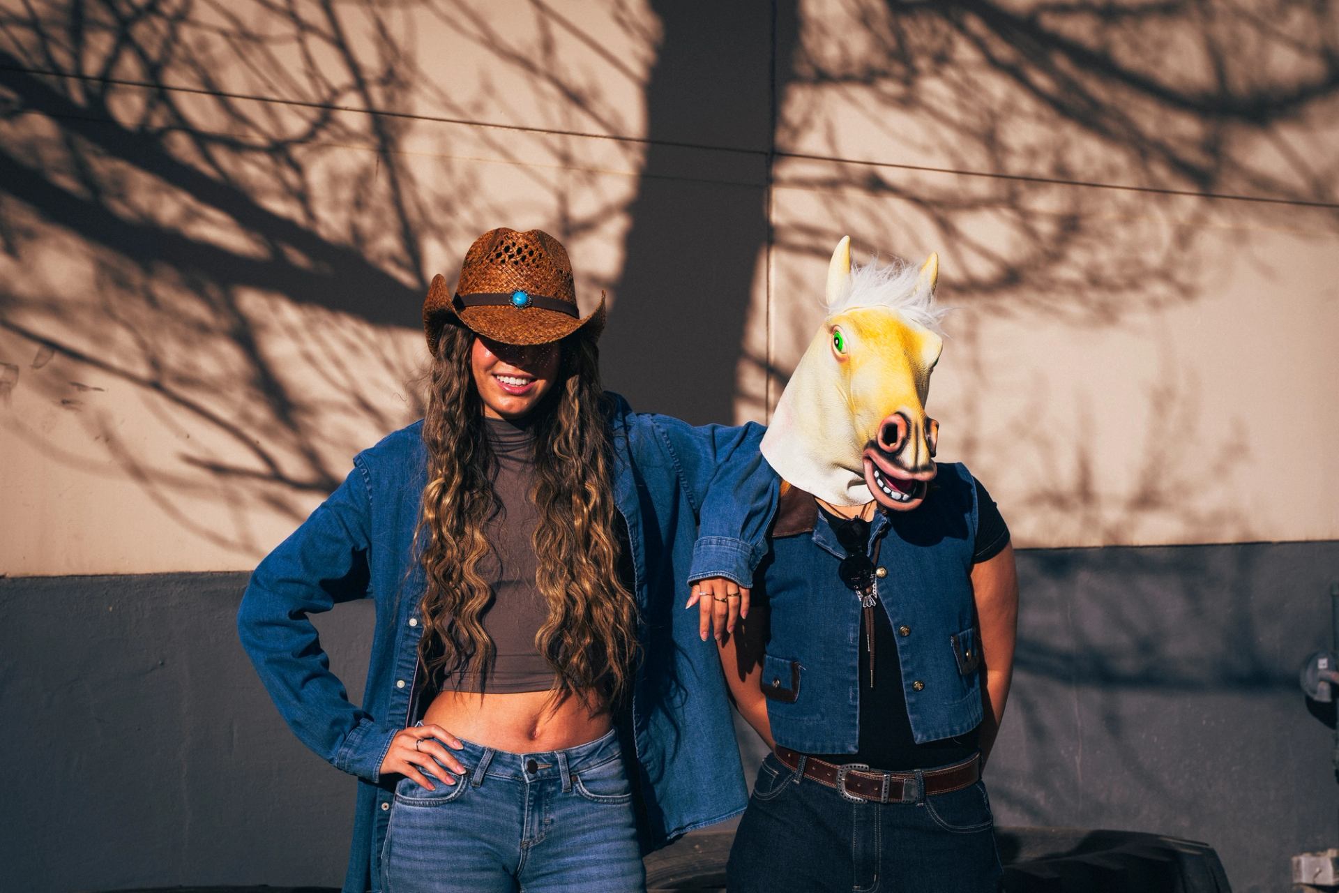 Two people in western outfits posing outdoors, one wearing a cowboy hat and the other a horse mask.