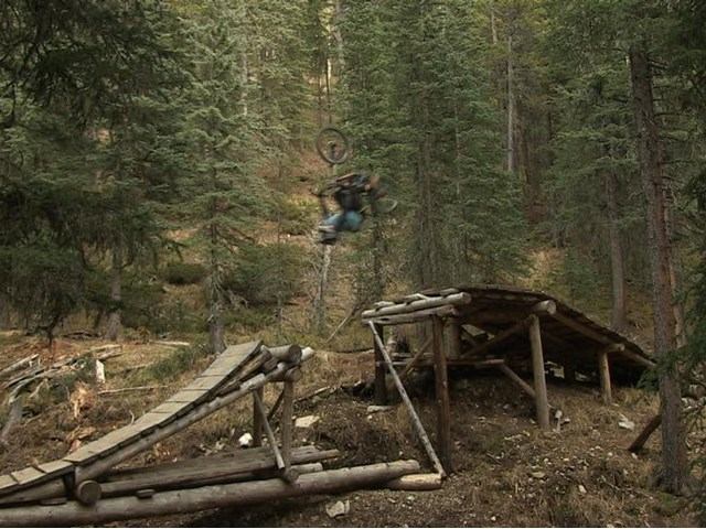 Mountain biker performing a jump off a wooden ramp in a dense forest.