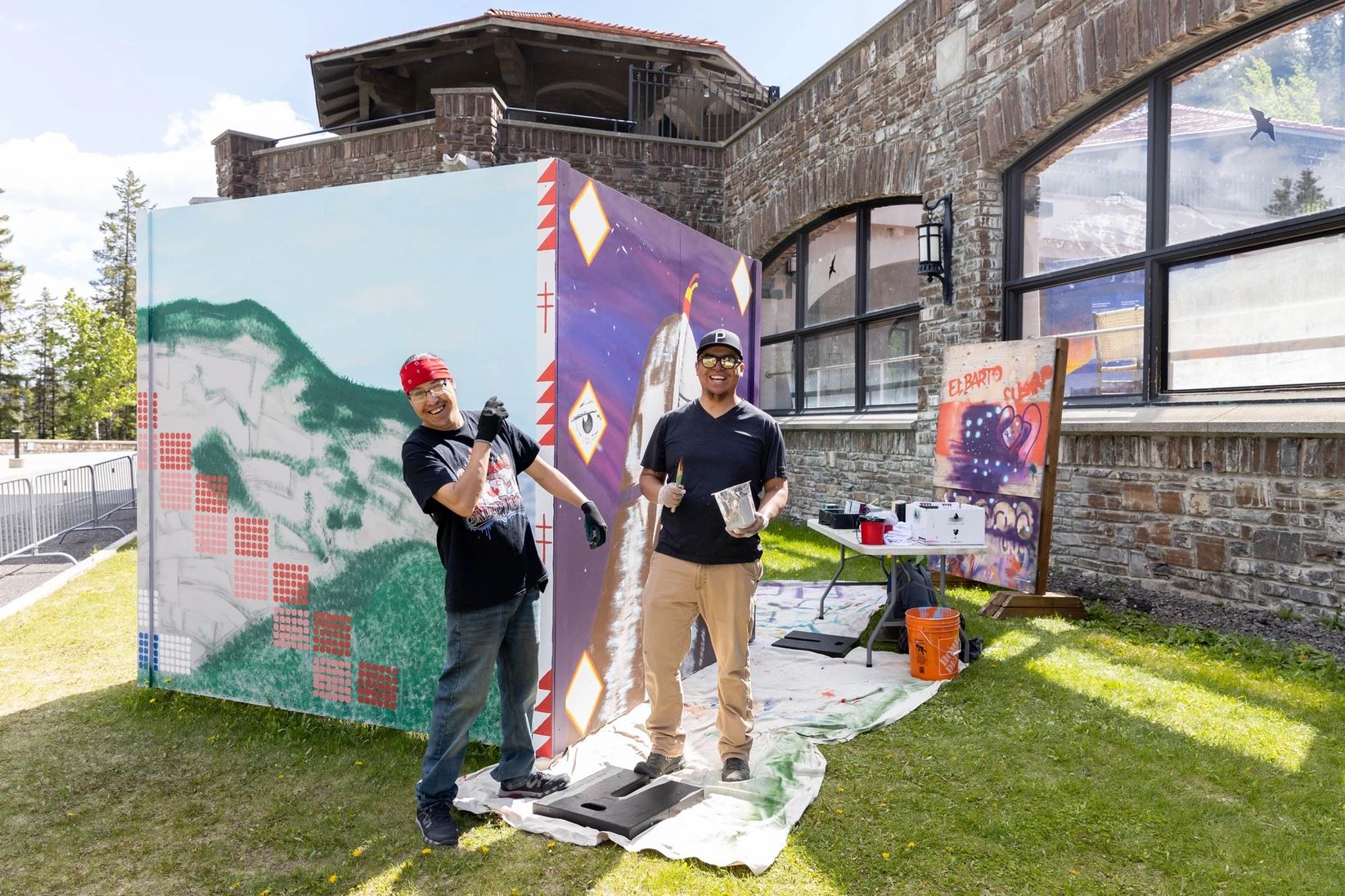 Artists in front of painted tipi backdrop, stone building, and supply table outdoors.