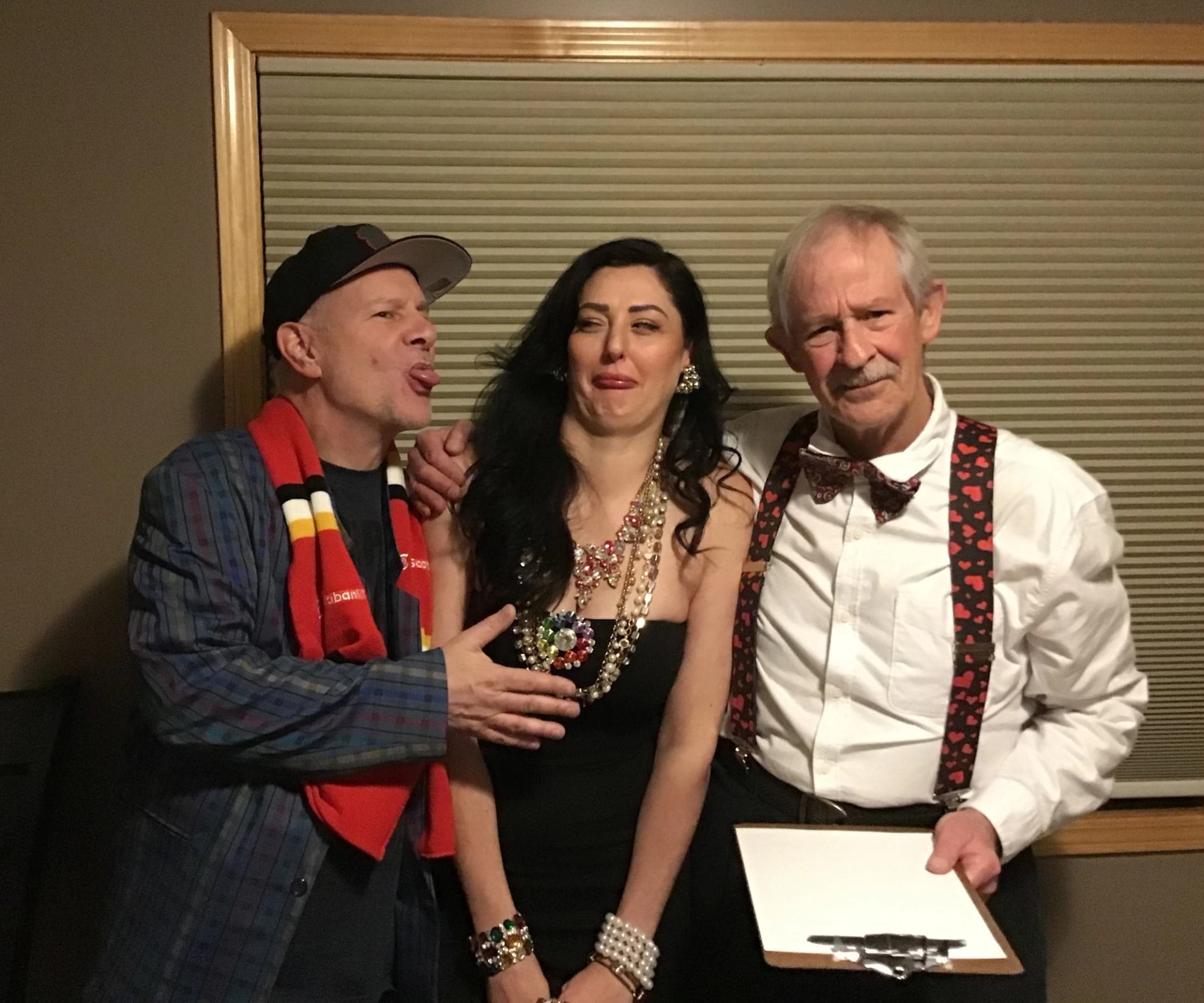 Three people pose in playful attire before a closed window blind.