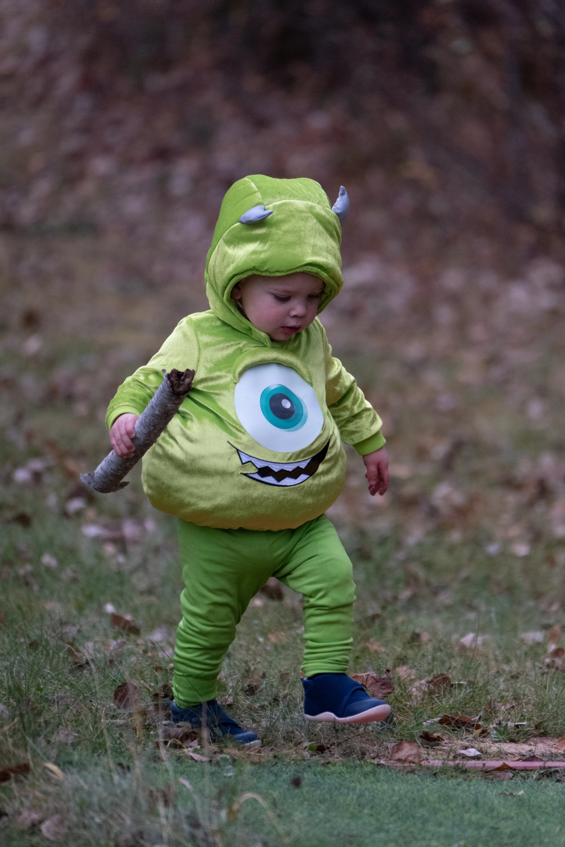 Child in green monster costume walks on grass while holding a stick outdoors.