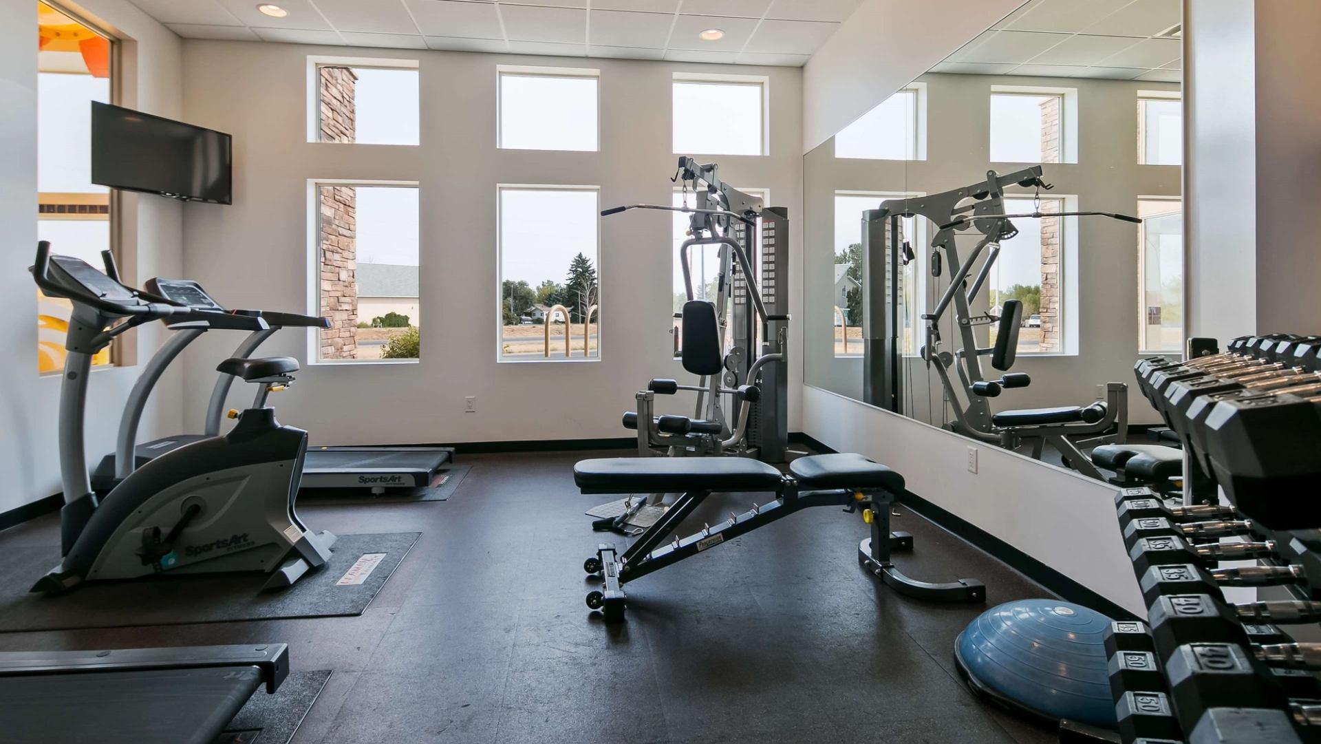 Hotel fitness room with treadmill, elliptical, weight bench, dumbbells, mirrors and windows.