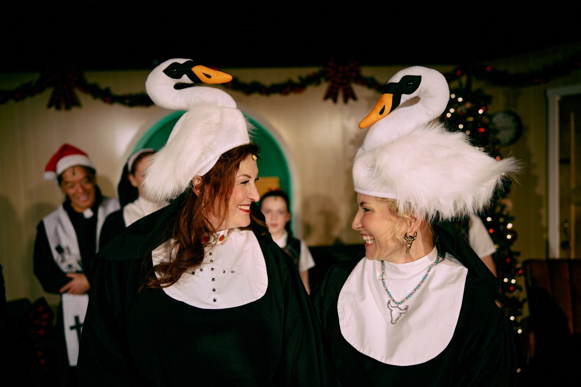 Two people in swan costumes face each other amid festive holiday decorations.