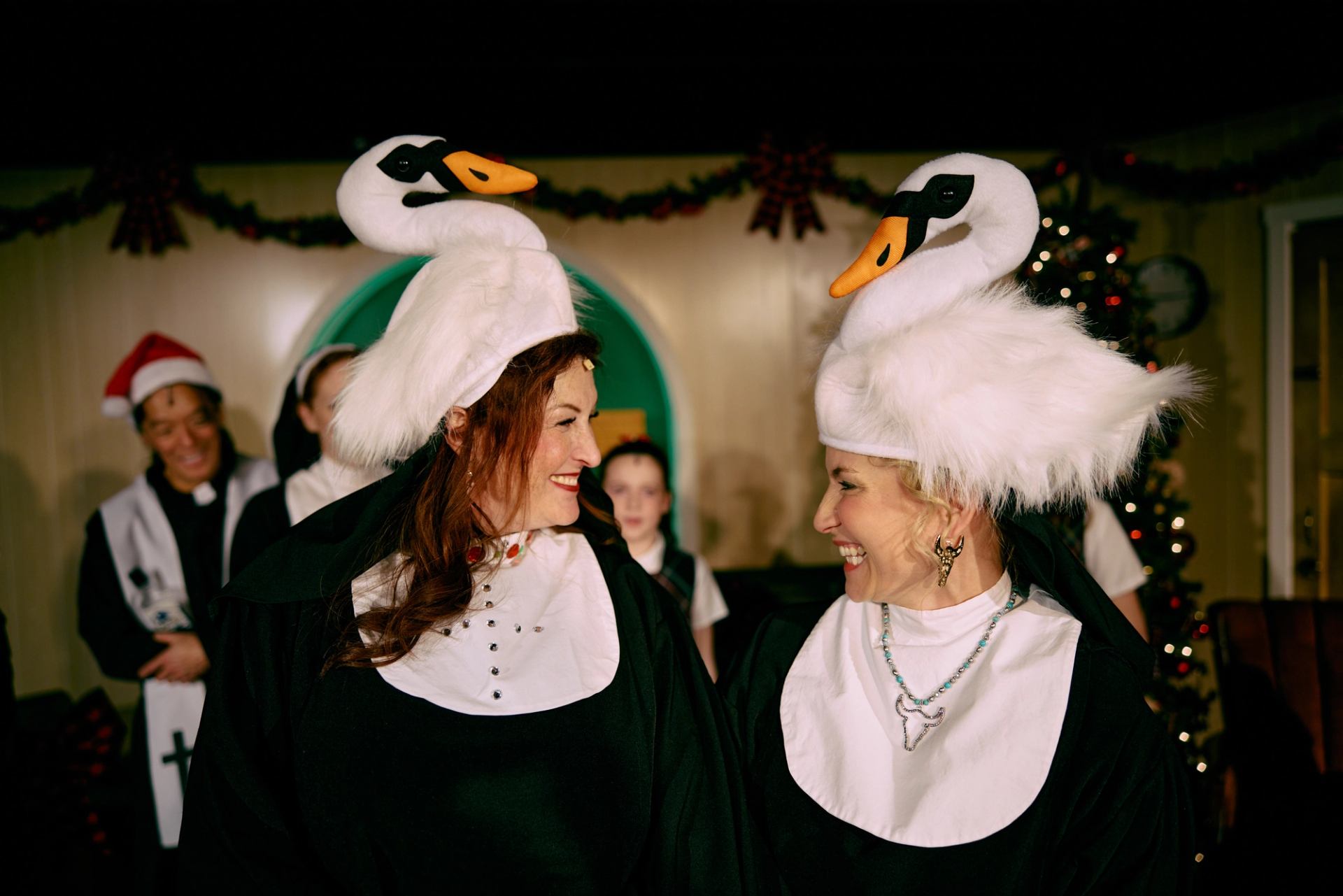 Two people in swan costumes face each other amid festive holiday decorations.
