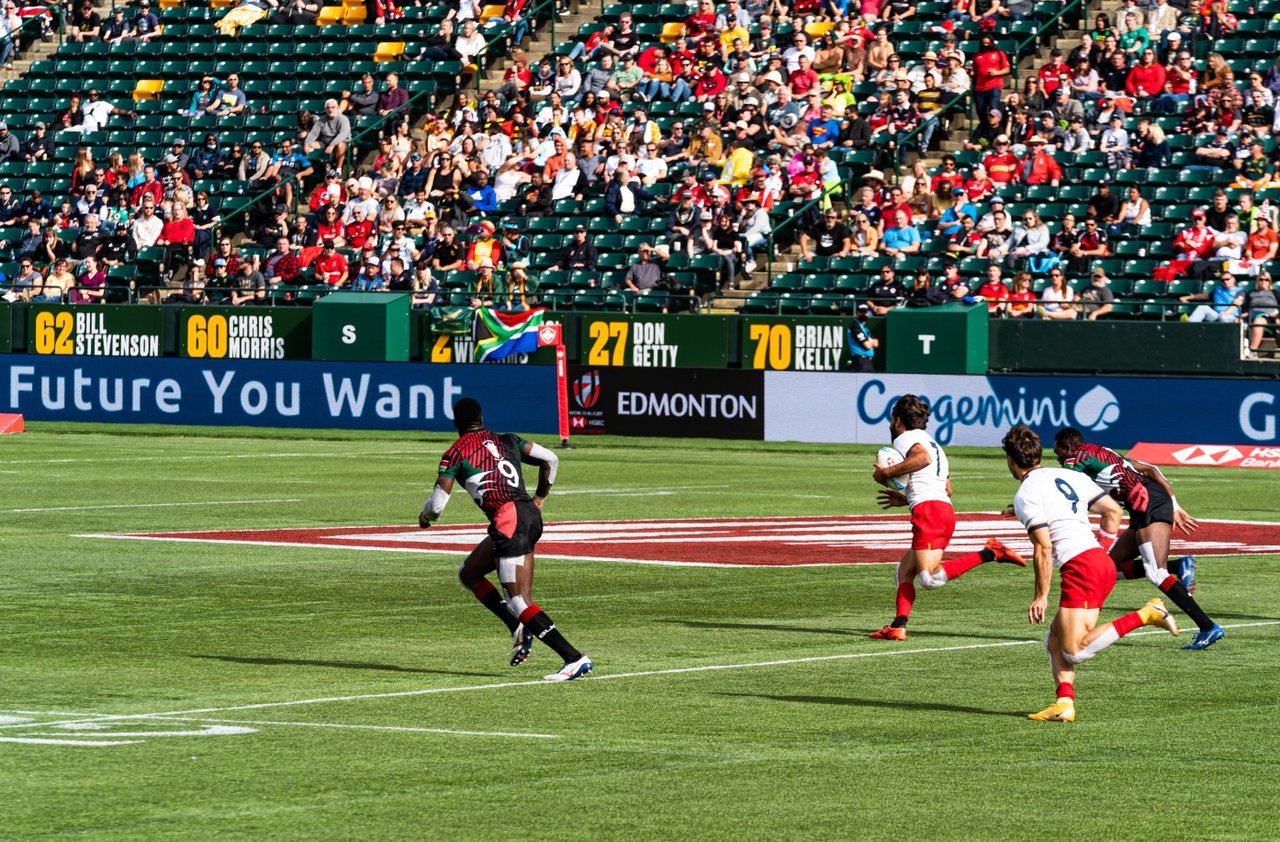 Rugby players sprint across stadium field as crowd watches from green bleachers.