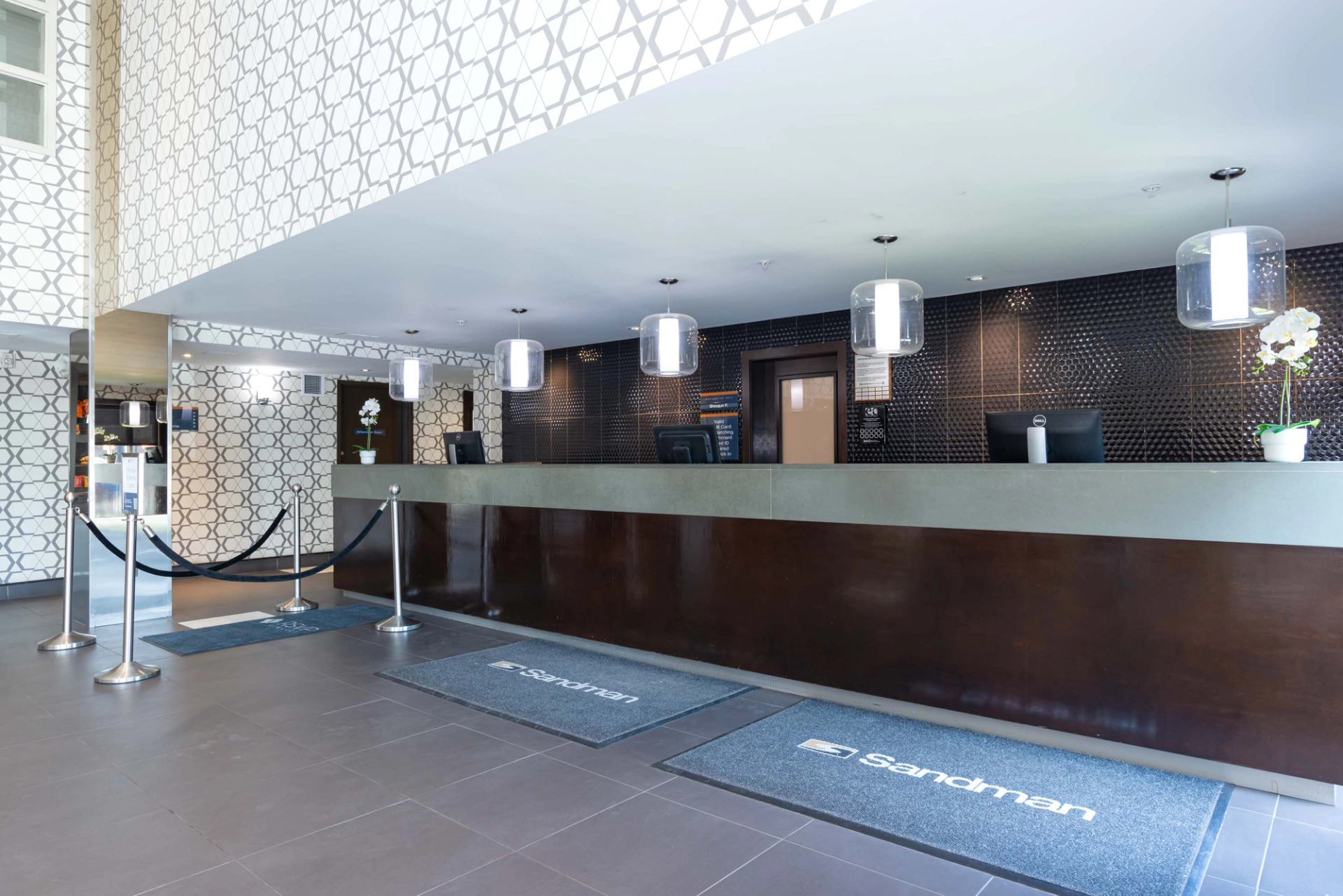 Hotel reception desk with patterned walls, pendant lights, Sandman logo mats and stanchions.