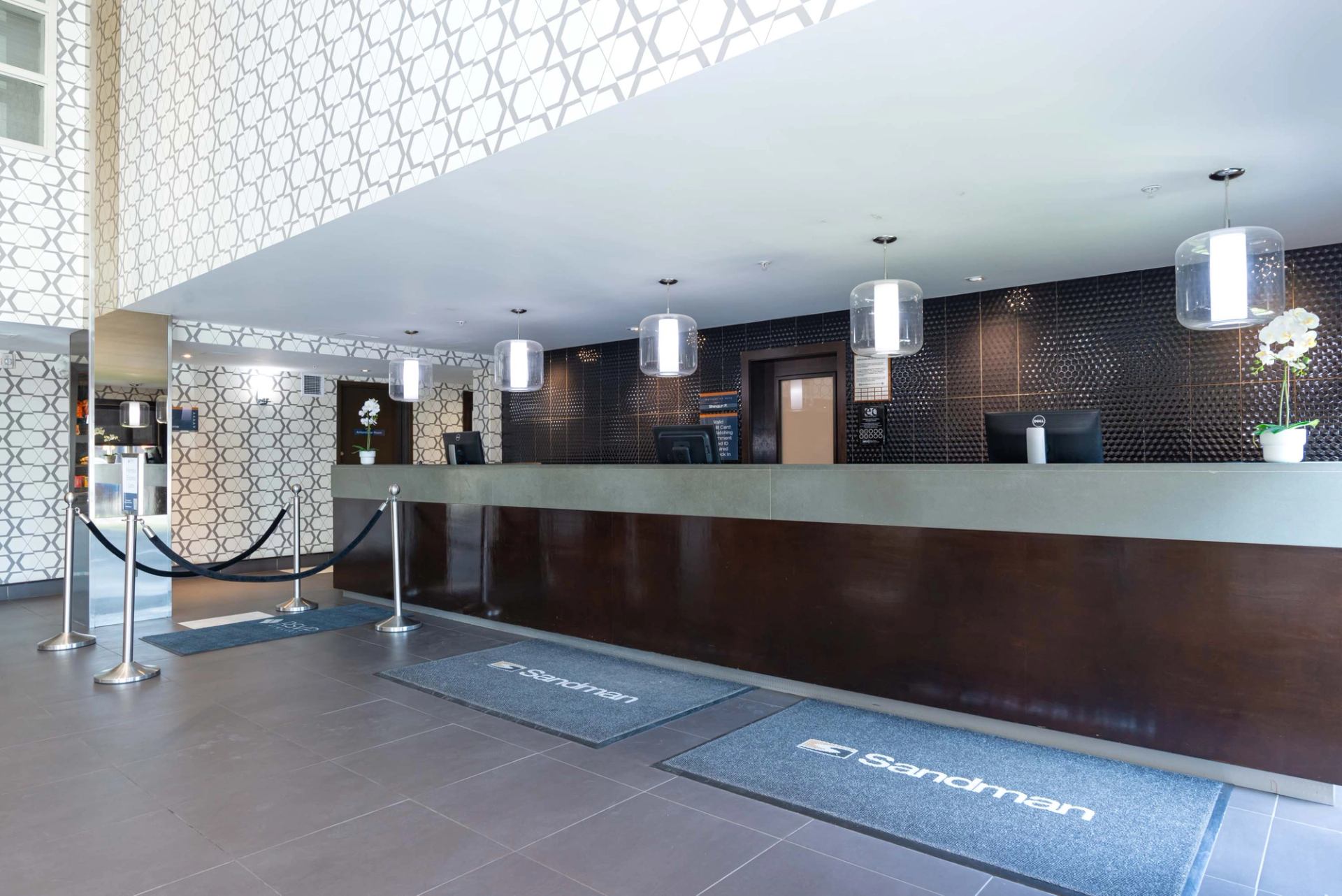 Hotel reception desk with patterned walls, pendant lights, Sandman logo mats and stanchions.