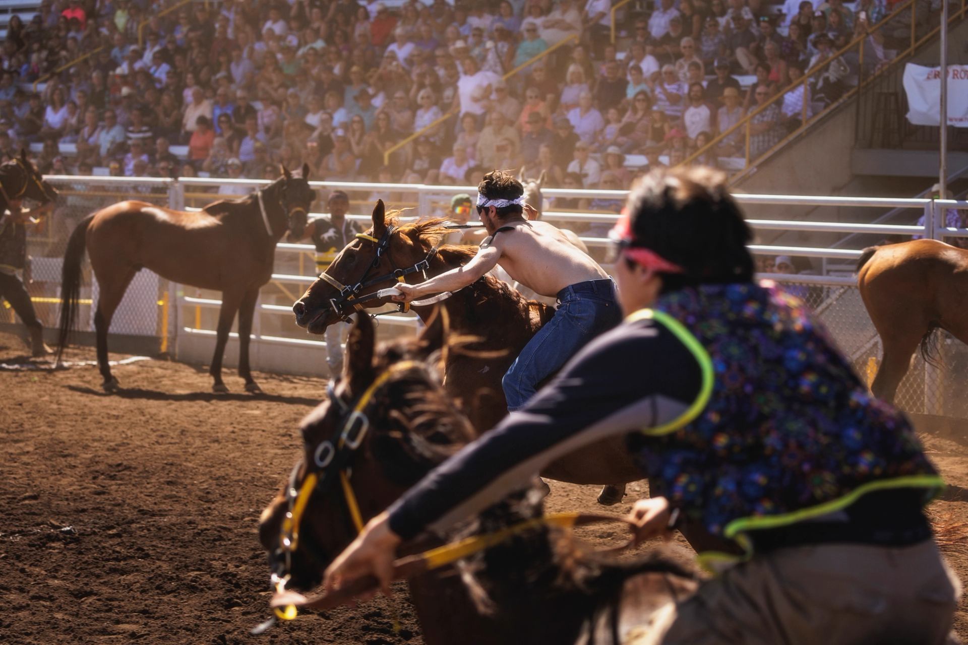 Bareback riders race horses on a dirt track before a large seated crowd.