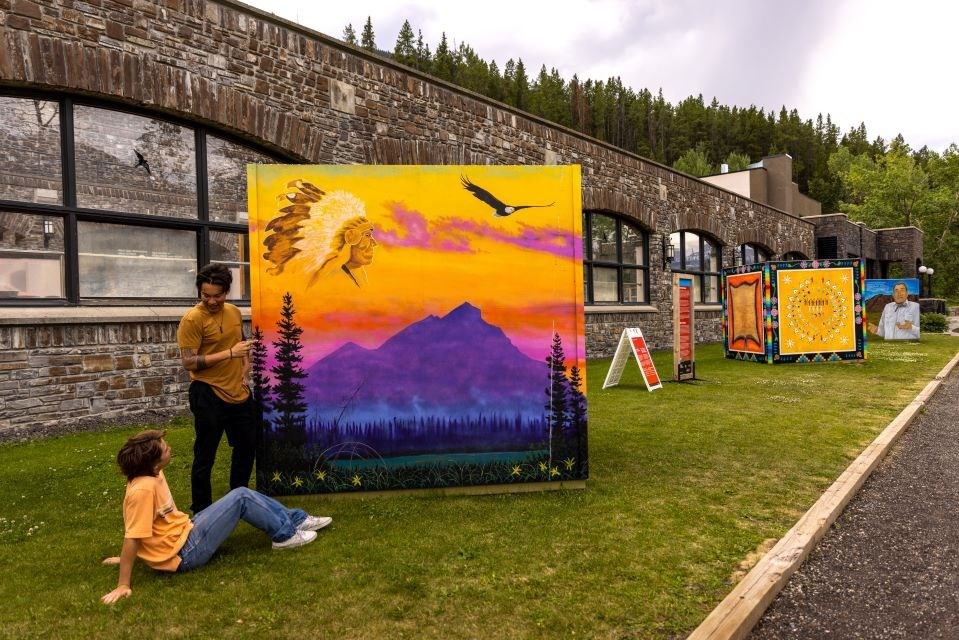 Outdoor art display with colourful Indigenous-style paintings and two people talking on the grass.