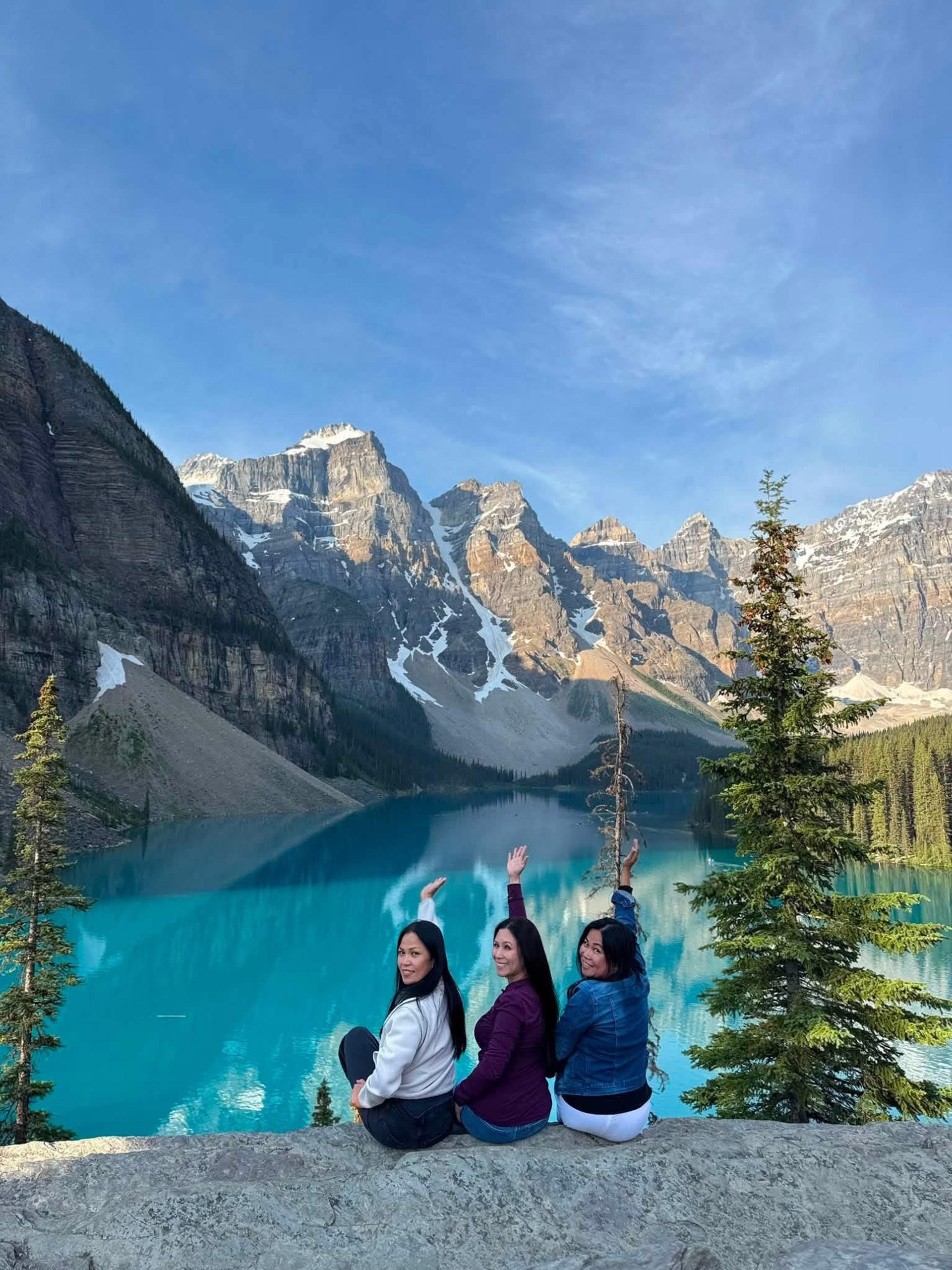 Three people sit above a turquoise mountain lake, raising their arms.