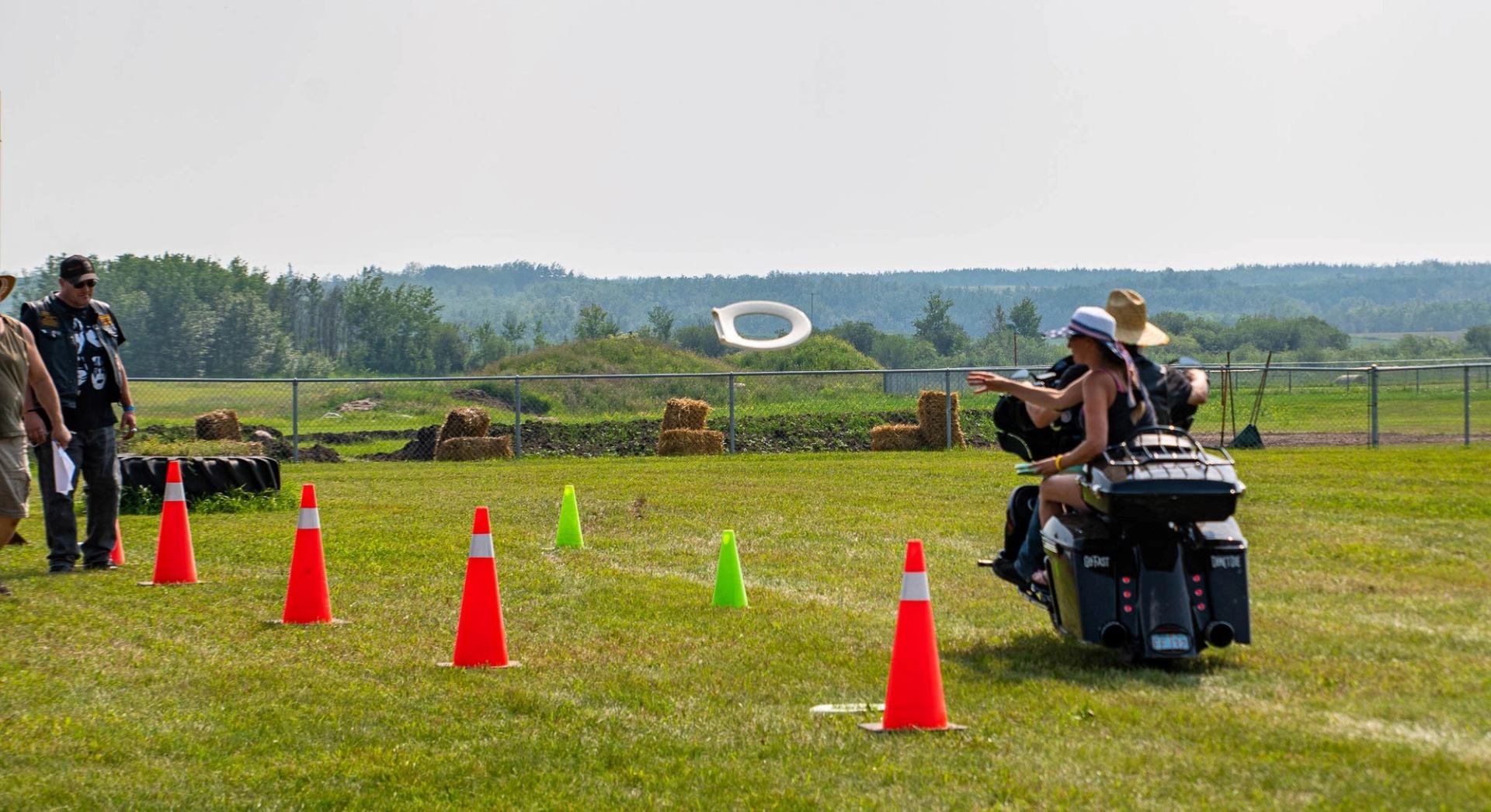 Motorcycle riders pass traffic cones on grass as a toilet seat flies nearby.