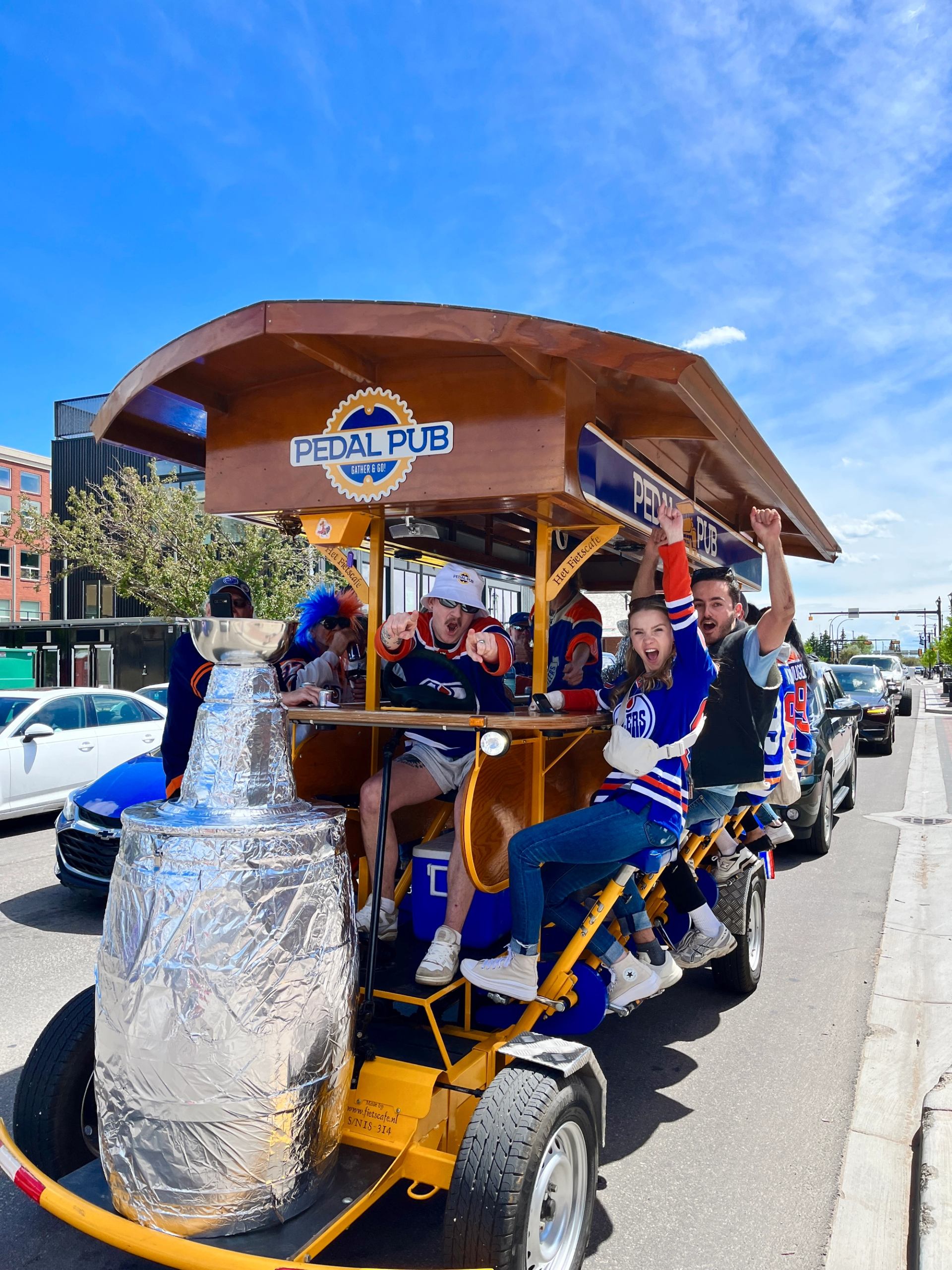 Fans celebrating on a Pedal Pub bike in Edmonton, riding downtown with team jerseys and raised arms.