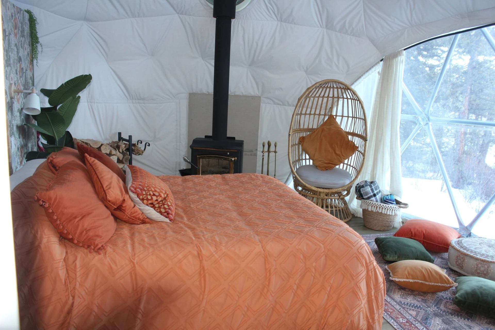 Bedroom in geodesic dome with orange bedding, woodstove, wicker chair and floor cushions.